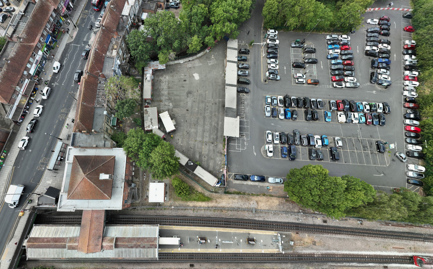 Aerial view of a railway station and surrounding area, showing station platforms and tracks in the foreground, an adjacent car park, and a cleared site compound between the station and town centre buildings. Mature trees border the site, with roads, parked vehicles and commercial properties visible around the perimeter.