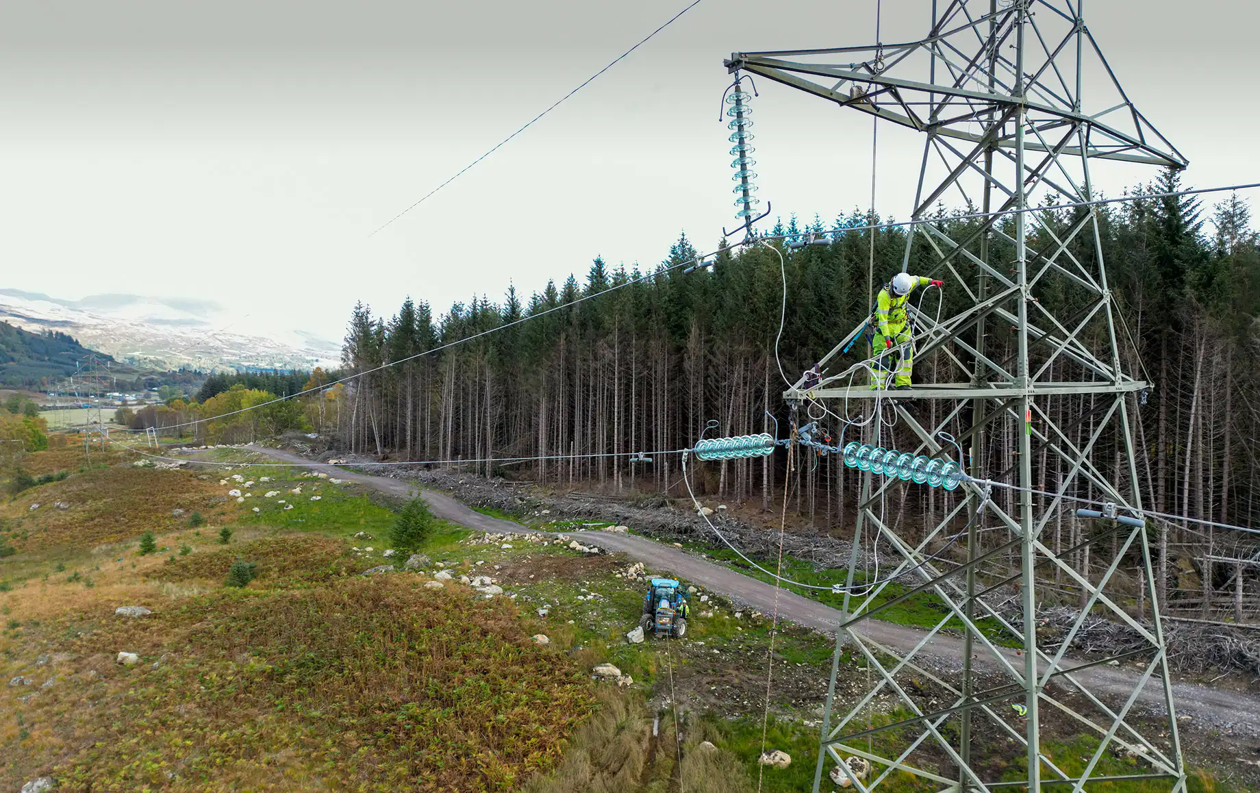 Worker in safety gear climbing a high-voltage transmission tower in a rural area, surrounded by forested hills, performing maintenance or installation on power lines.