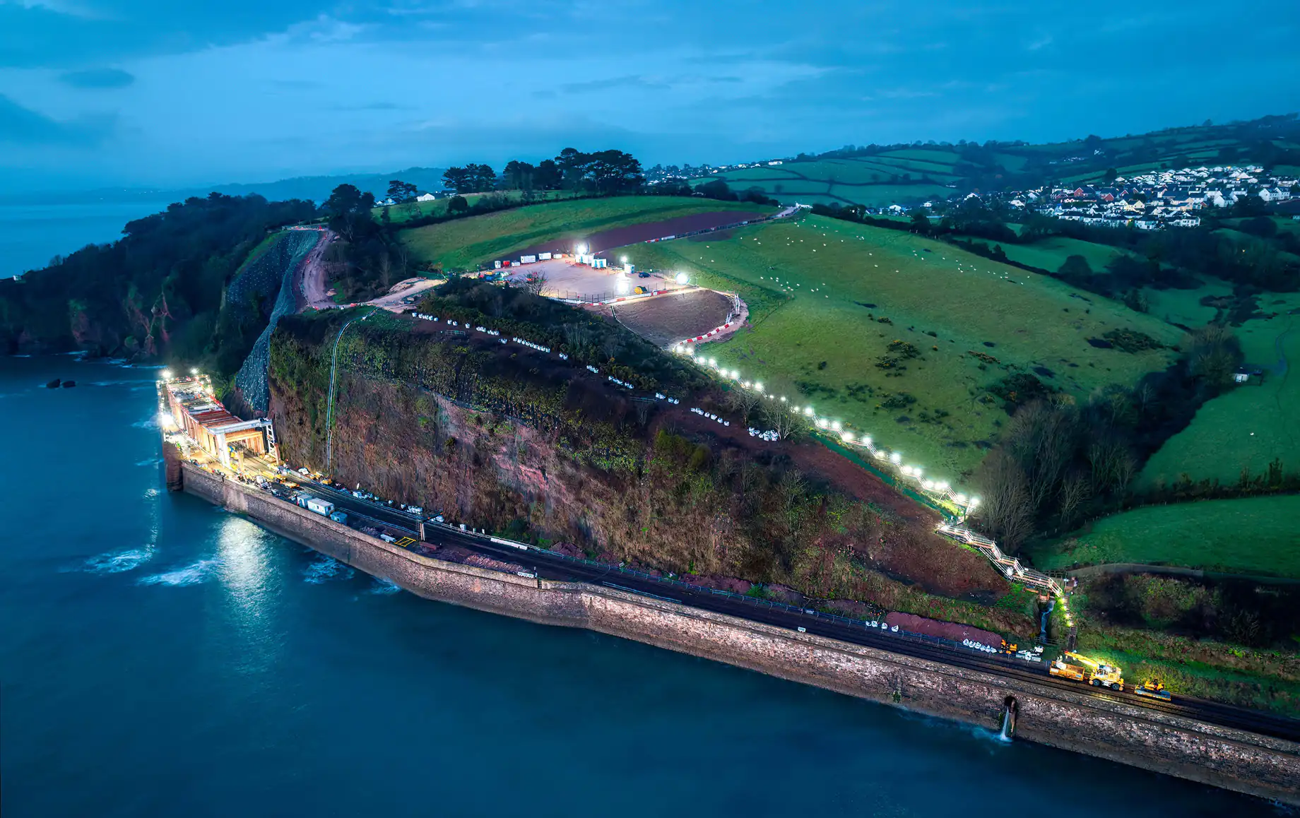 Aerial view of a coastal construction site at dusk, showing illuminated pathways and equipment along a cliffside, with green fields and residential buildings in the background.