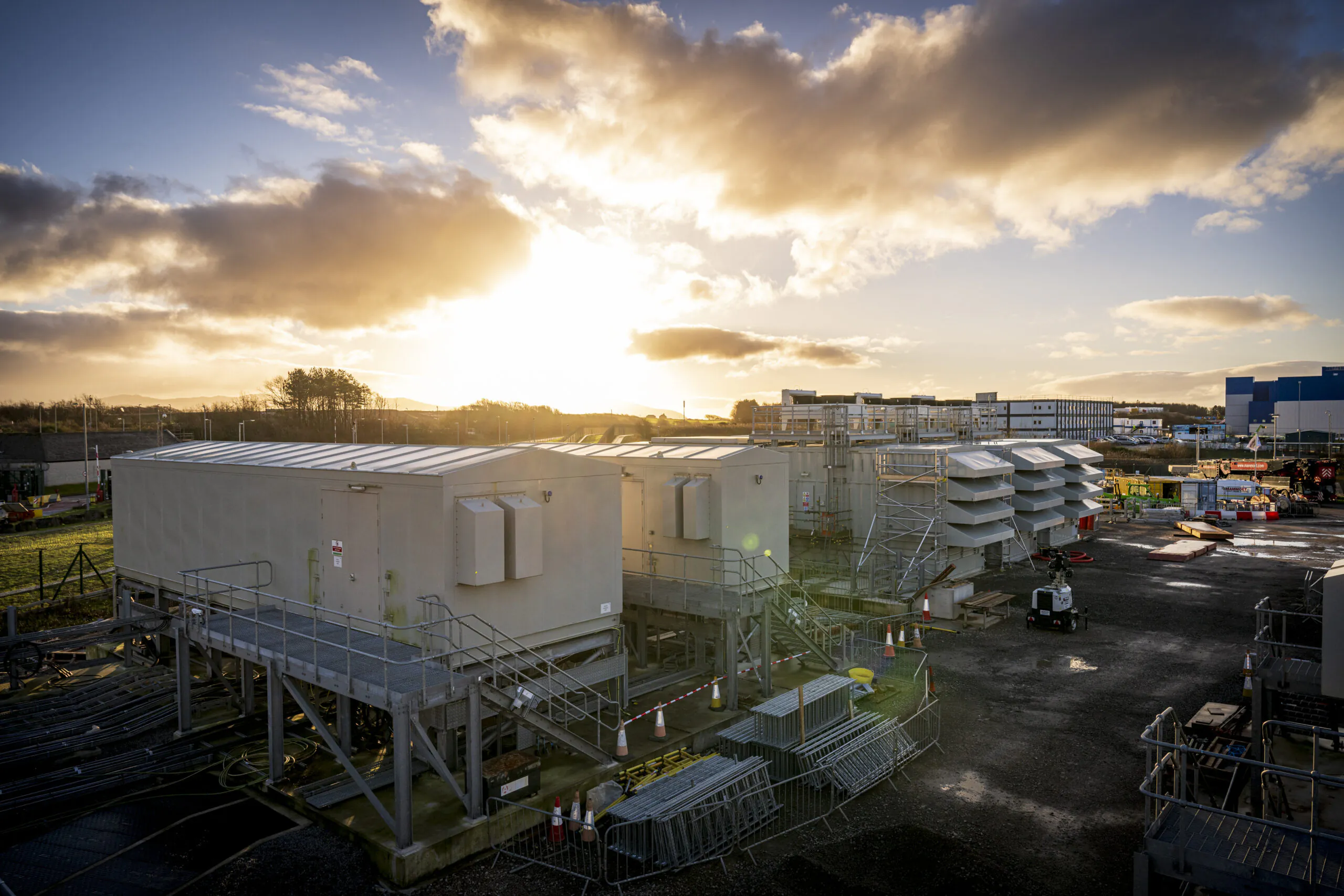 Large industrial site at sunset with modular buildings elevated on metal platforms, surrounded by construction equipment and materials.