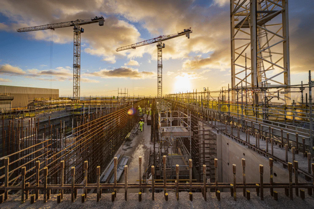 Wide-angle view of a large construction site at sunset with cranes, scaffolding, rebar structures, and workers across multiple levels.