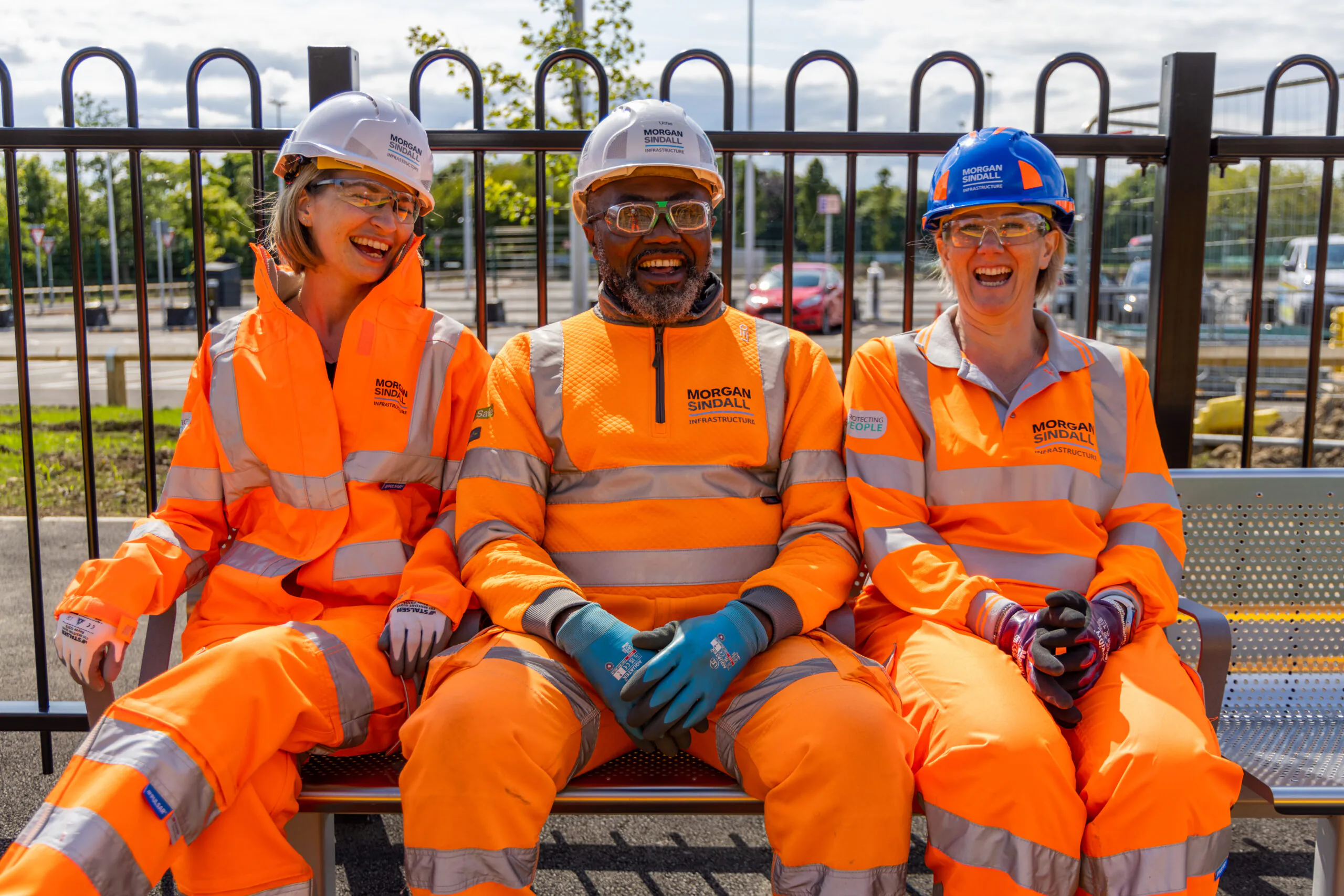 Three people in high-visibility orange clothing sitting on a bench outdoors and smiling.