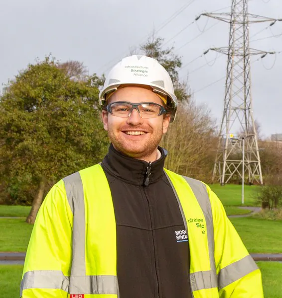 Person in high-visibility clothing and hard hat labeled 'Infrastructure Strategic Alliance' standing near power lines outdoors.