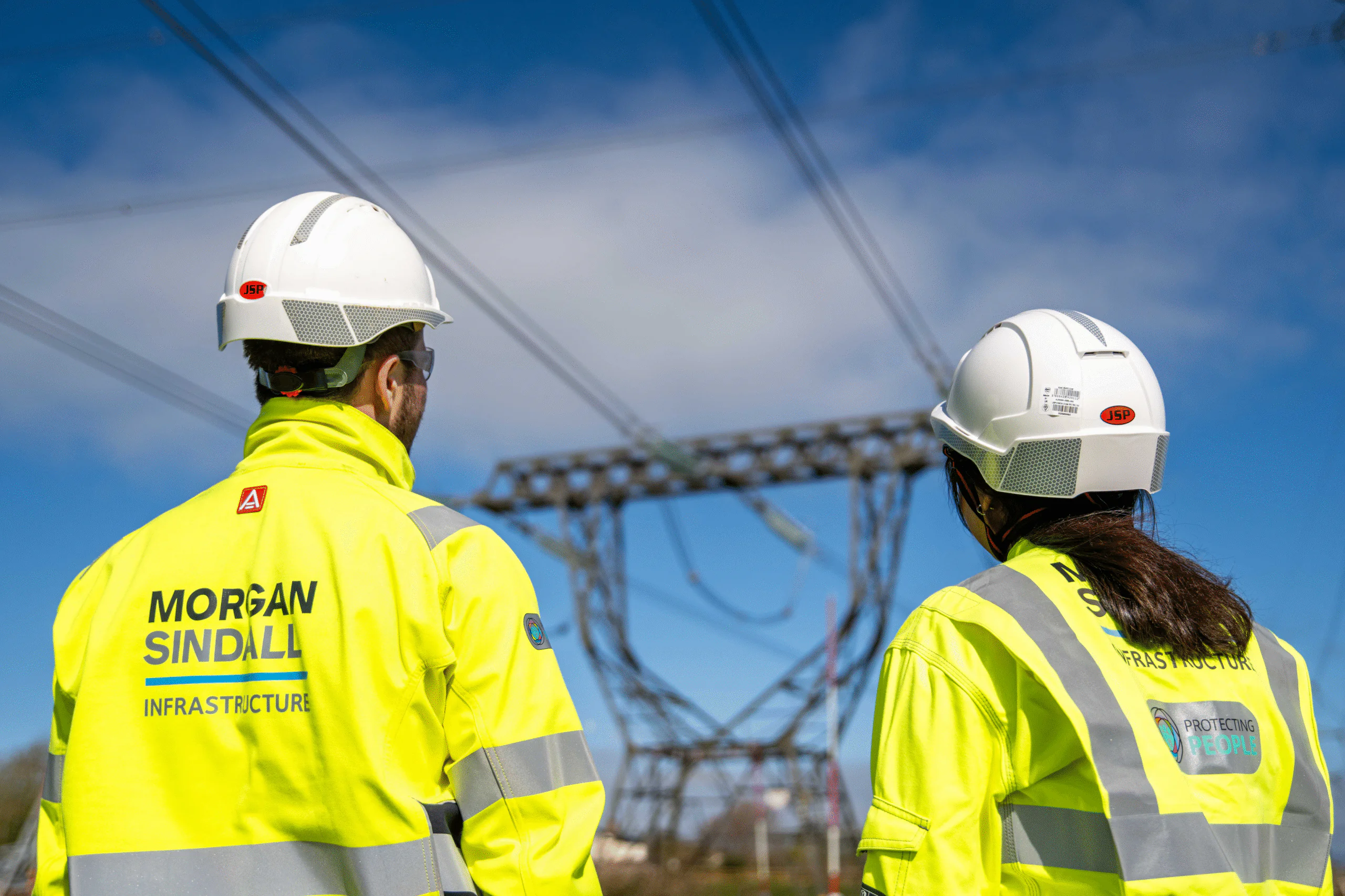 Two individuals in high-visibility jackets and white hard hats labeled 'Morgan Sindall Infrastructure' inspecting electrical infrastructure outdoors.