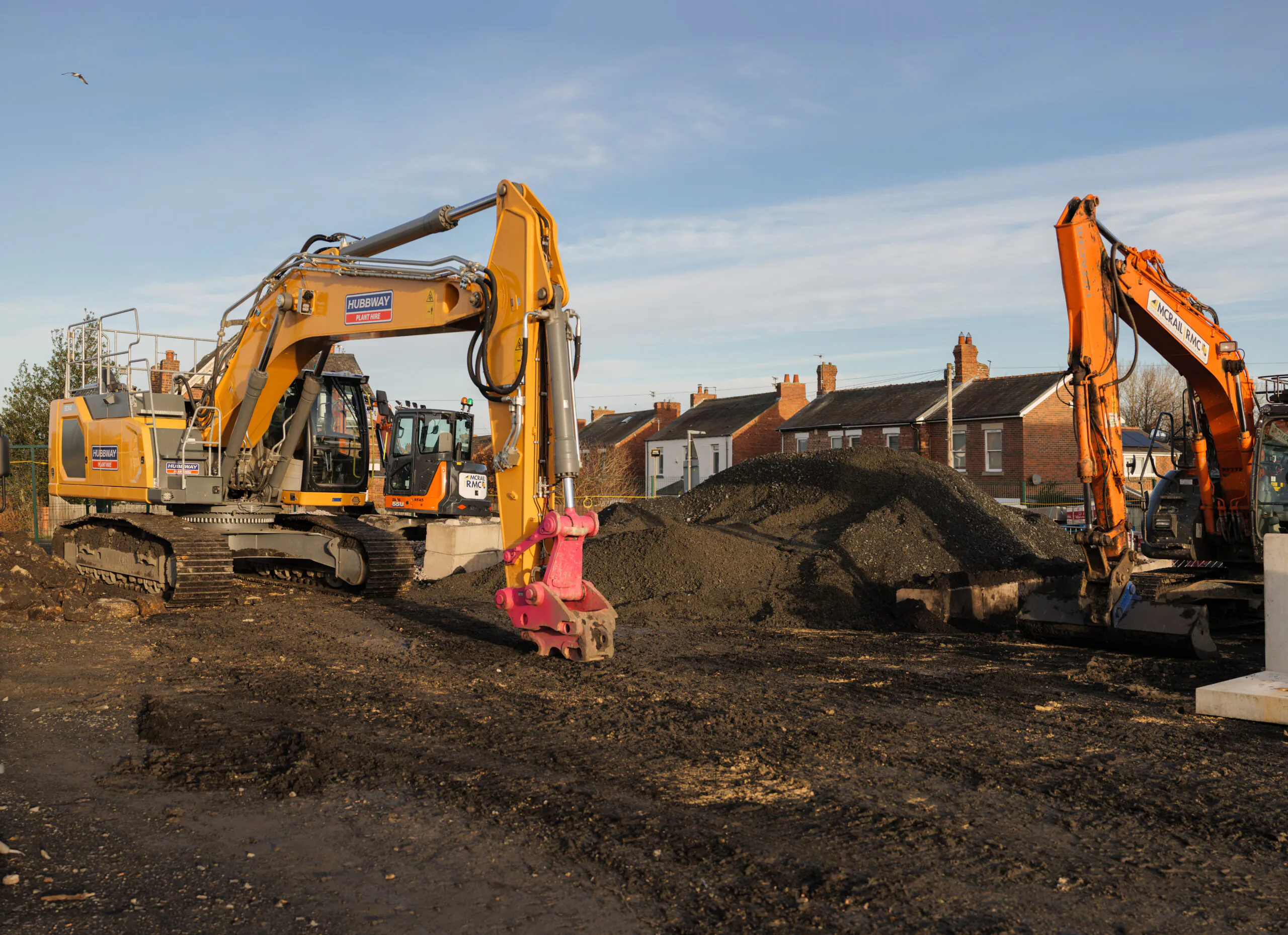 Close-up of two excavators working at a construction site with piles of dirt and residential houses in the background.