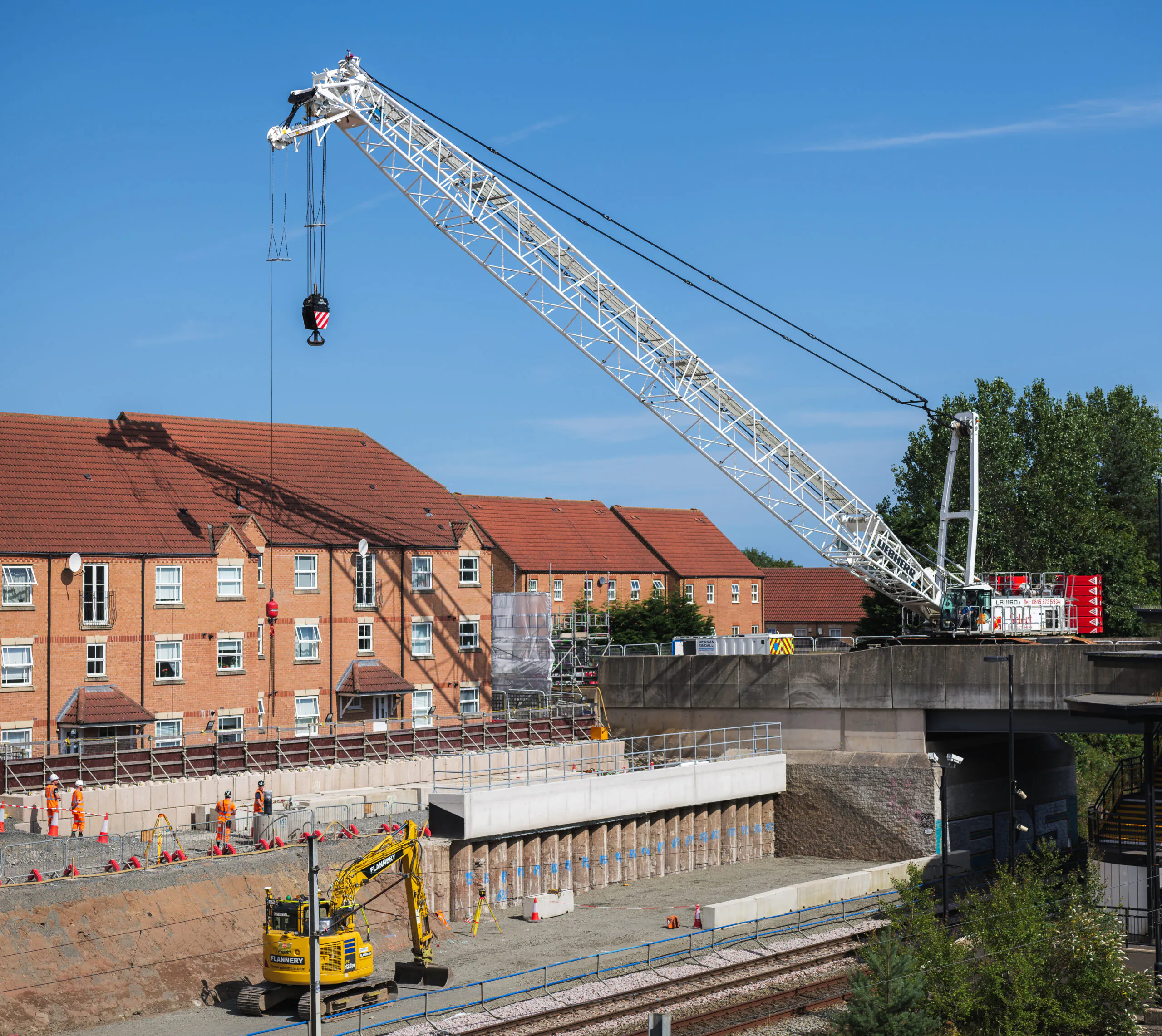 Daytime construction site with a large crane lifting materials near residential buildings and railway tracks.