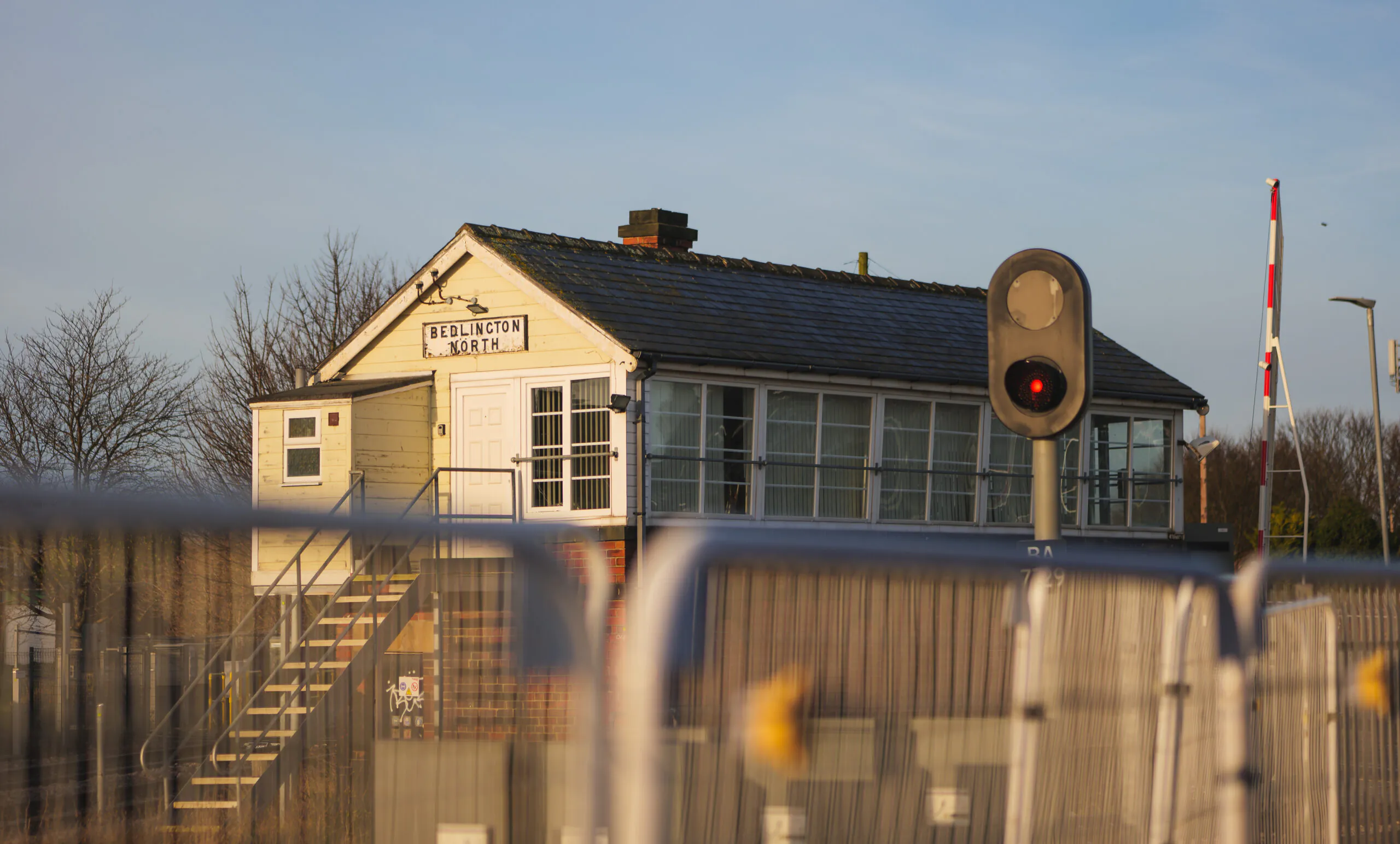 "Small brick building labeled 'Bedlington North' next to railway tracks, viewed through metal fencing with signal lights nearby.