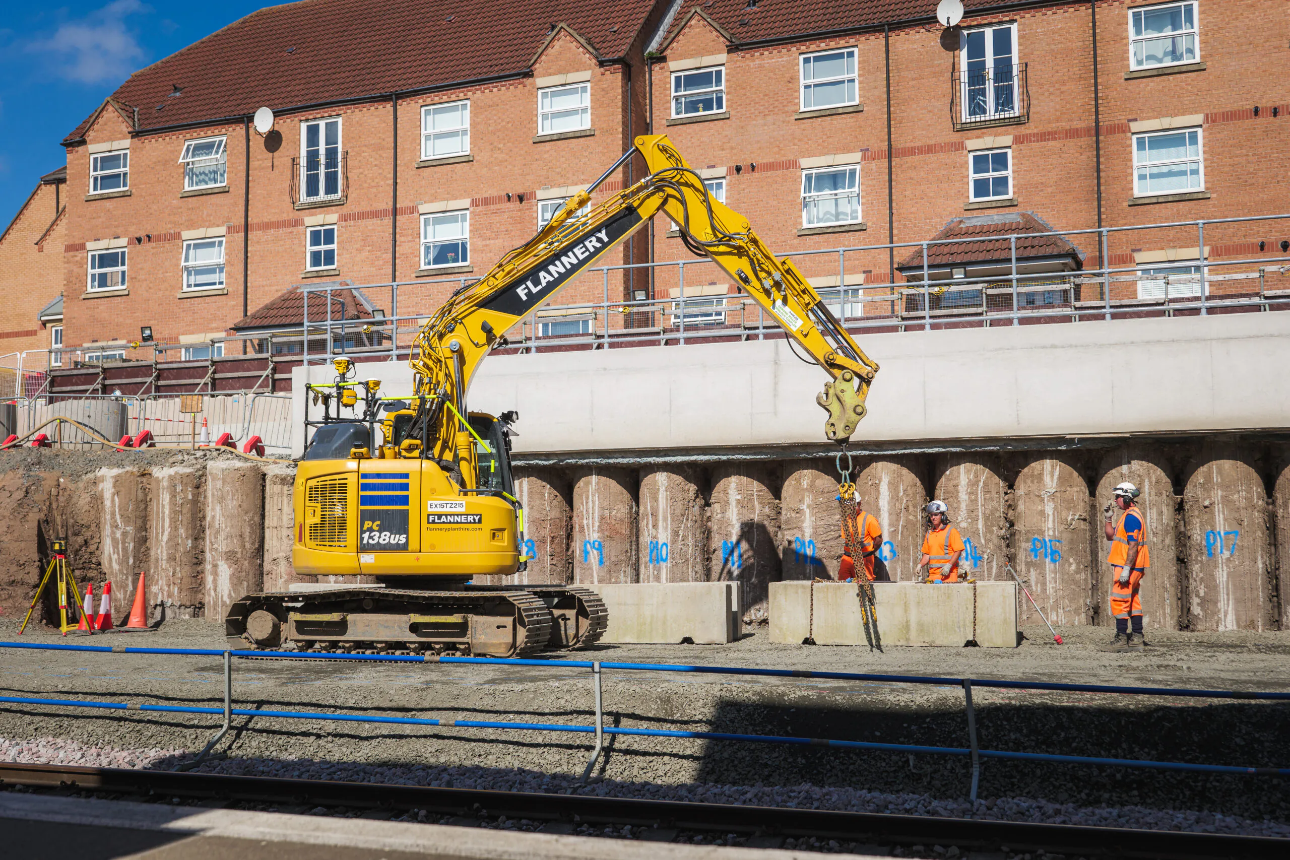 Excavator digging beside railway tracks during piling works, with temporary barriers and safety personnel present.