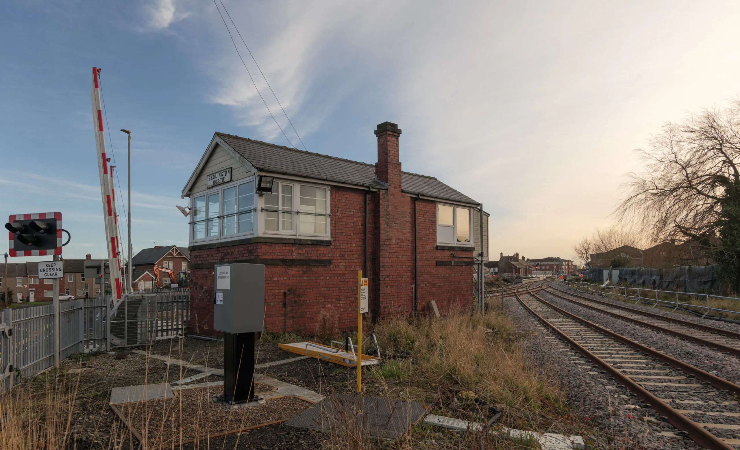 Small brick building next to railway tracks, surrounded by overgrown vegetation and fencing.