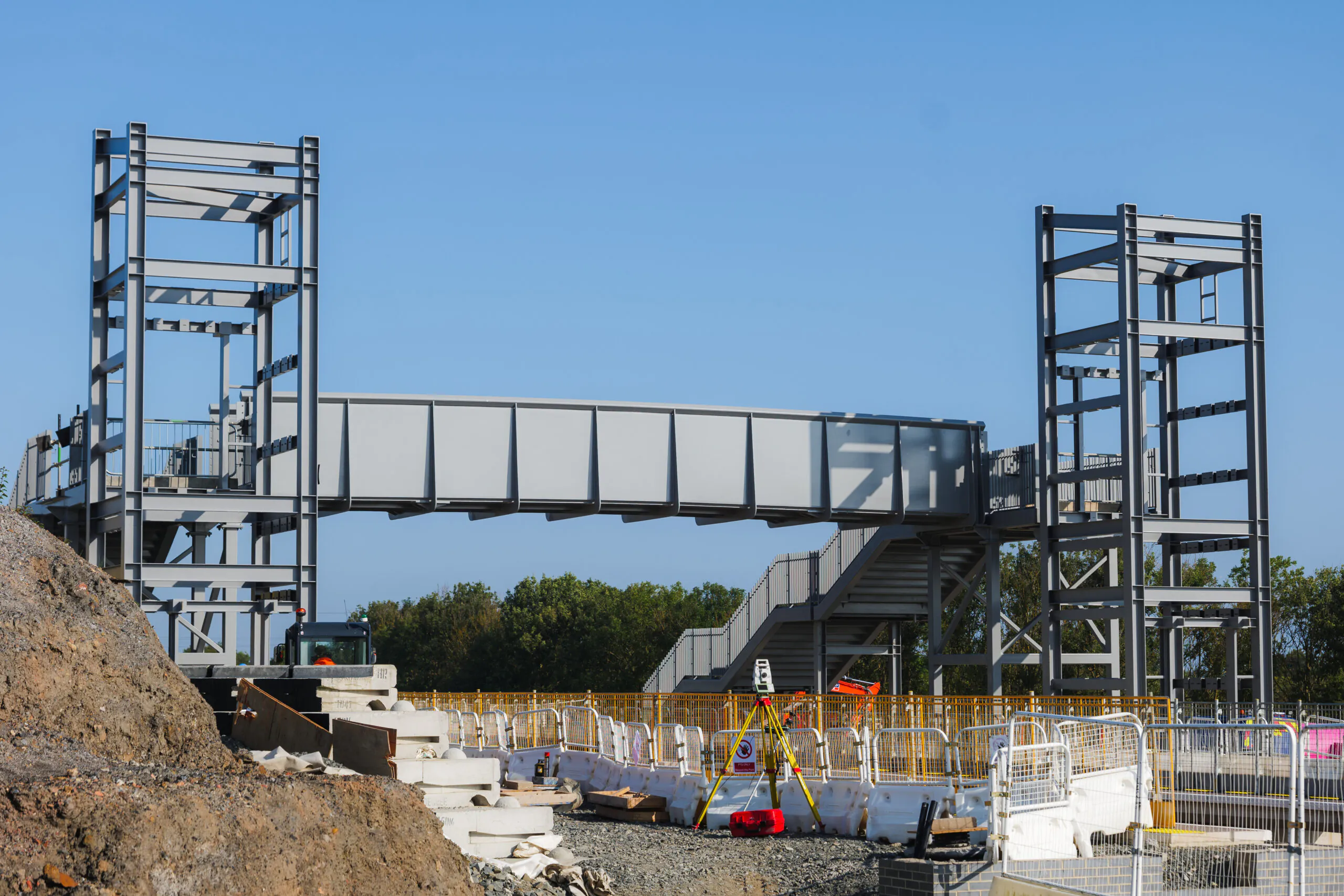 Partially constructed pedestrian bridge made of steel beams at an outdoor construction site under clear skies.