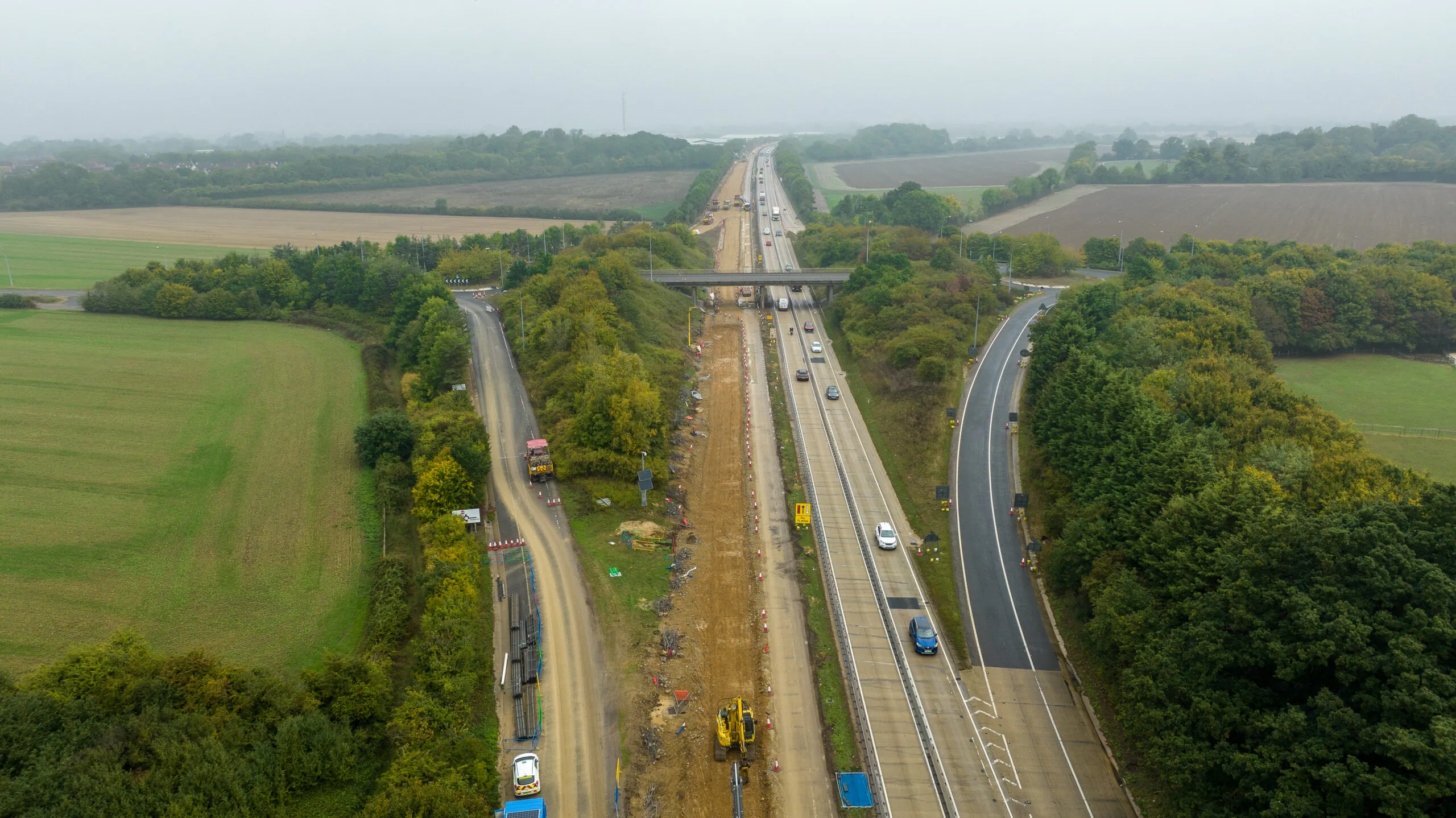 Aerial view of highway construction site with multiple lanes, vehicles, and equipment surrounded by fields and trees.