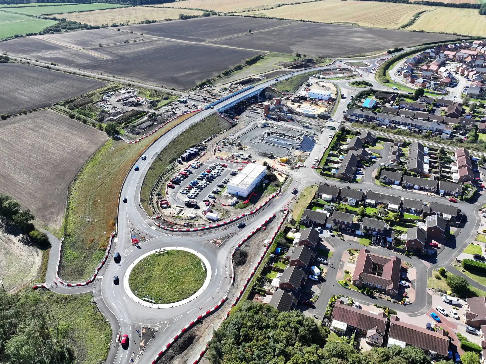 Aerial view of construction near residential areas with roads, roundabouts, buildings under development, and surrounding fields.