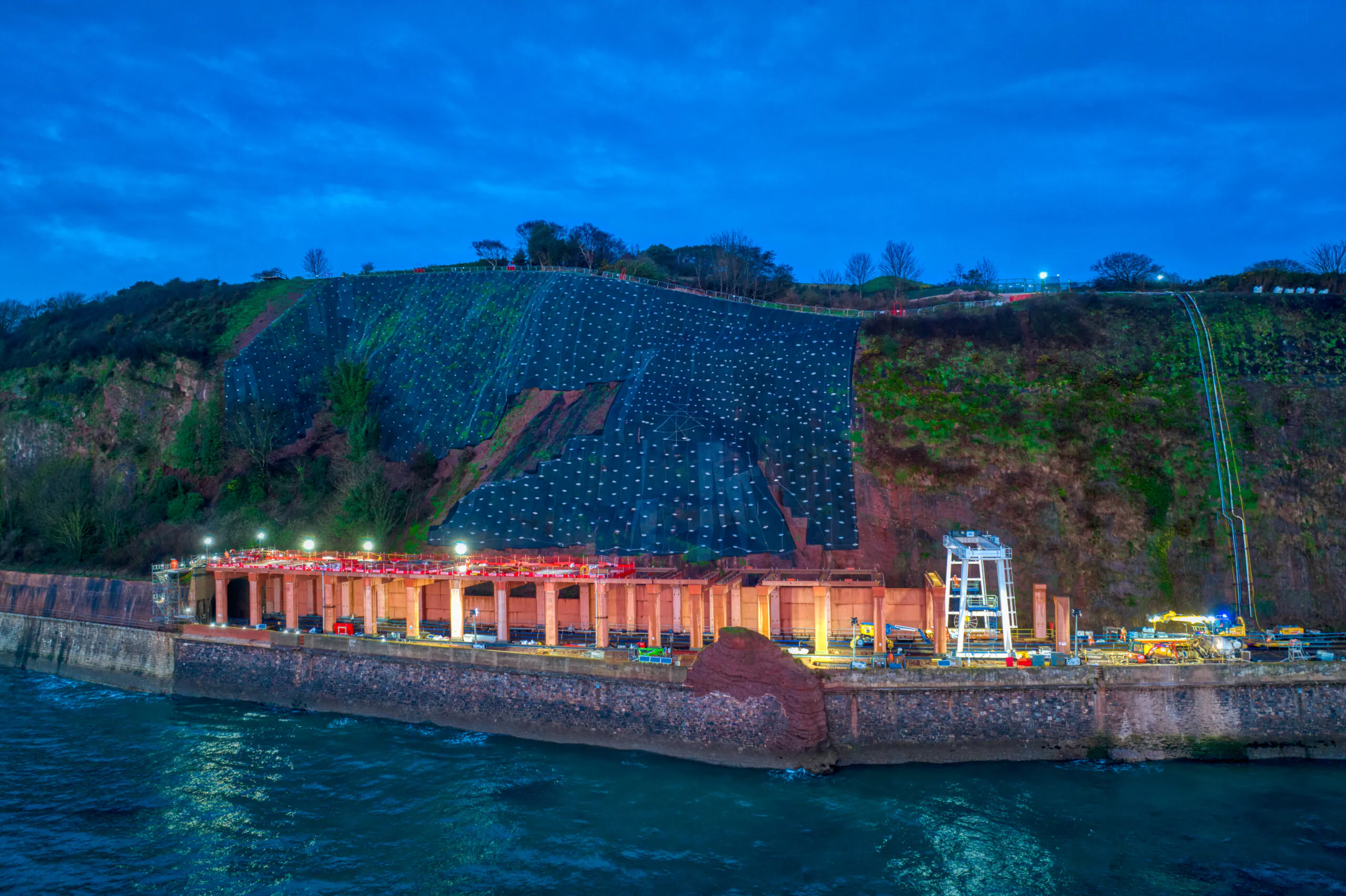 Wide-angle view of coastal railway construction site with cliffside stabilization and lighting setup at dusk.