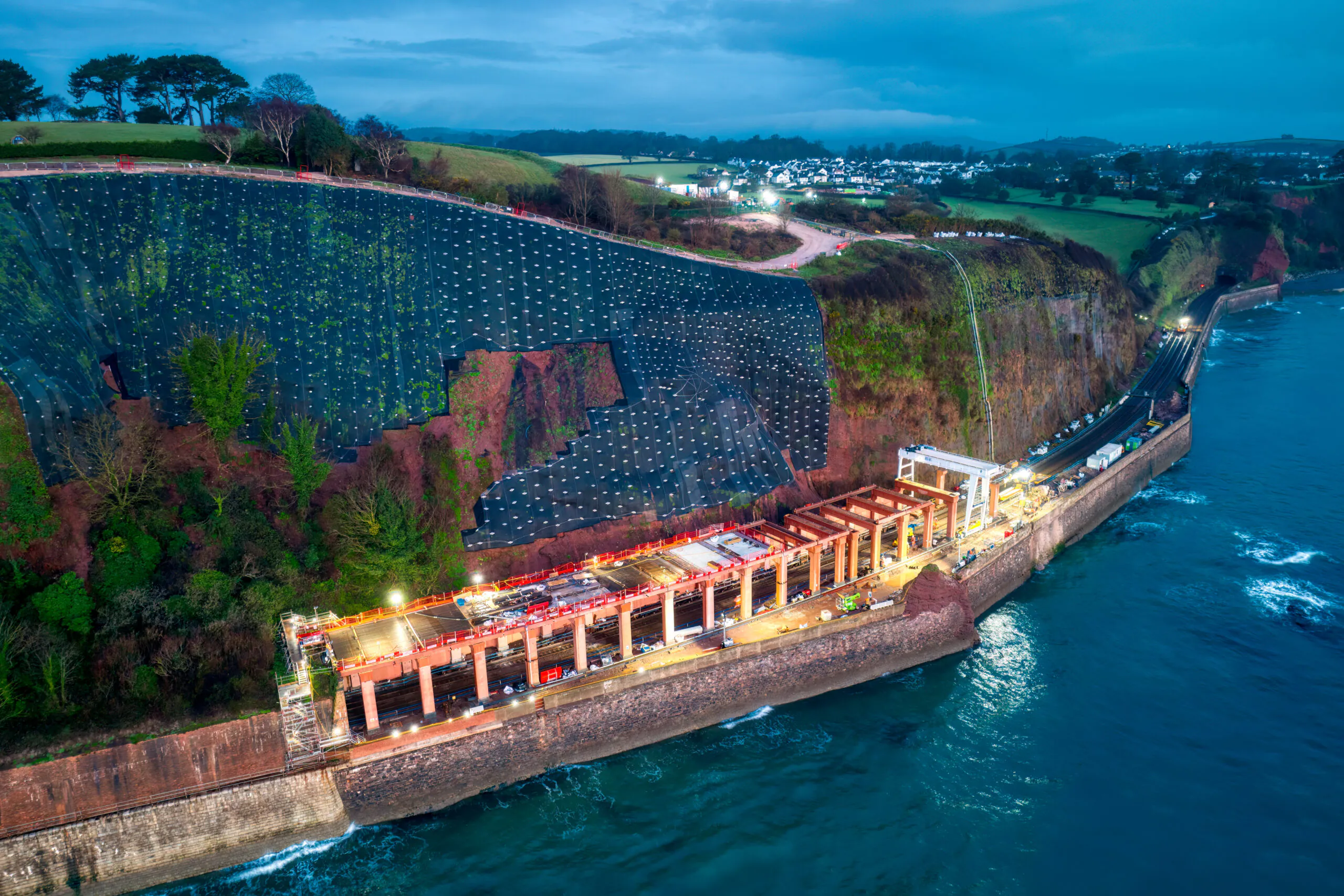 Aerial view of coastal construction work on a cliffside next to the sea, with lights illuminating the site.