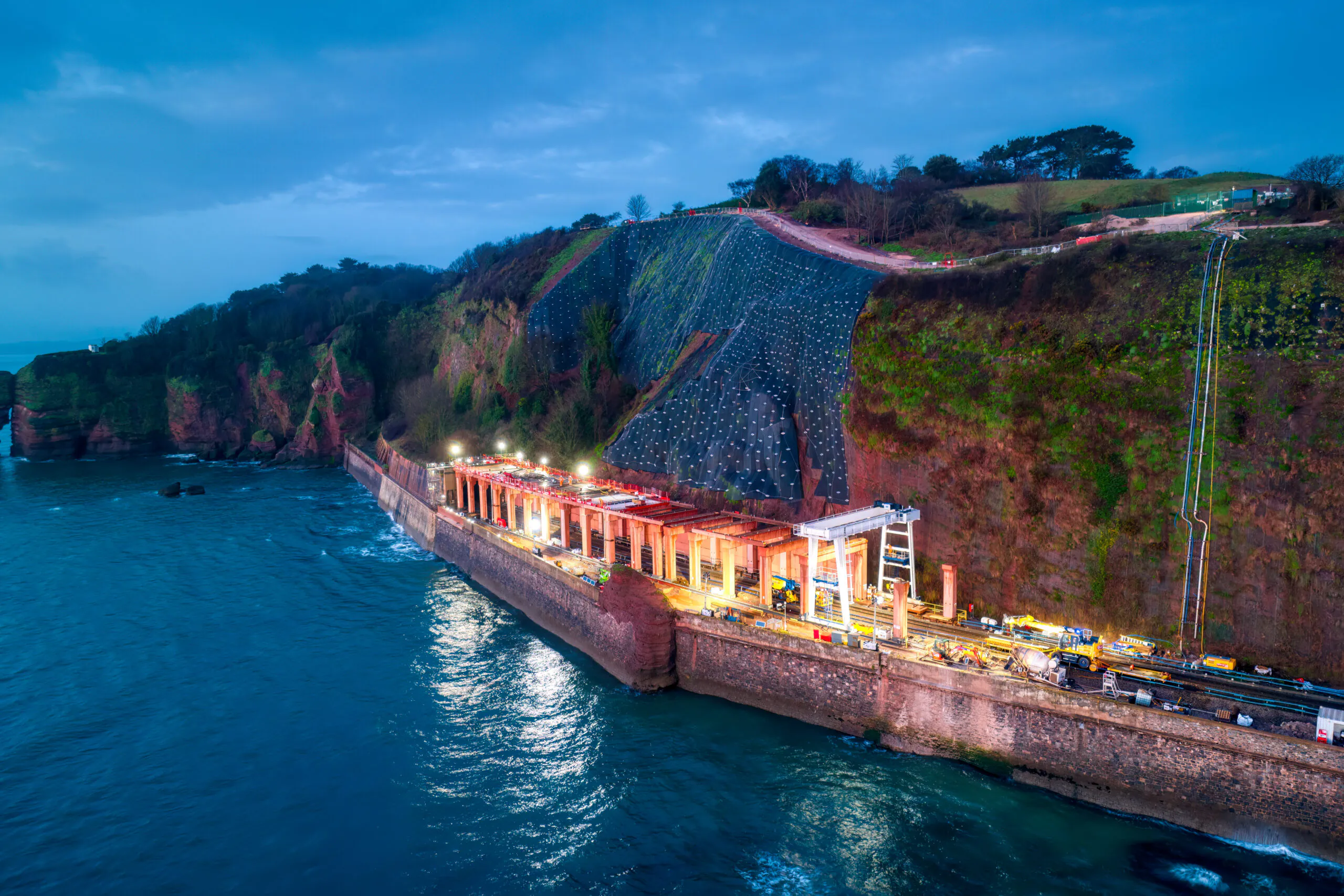 Nighttime construction work along a coastal railway line next to cliffs illuminated by bright lights.