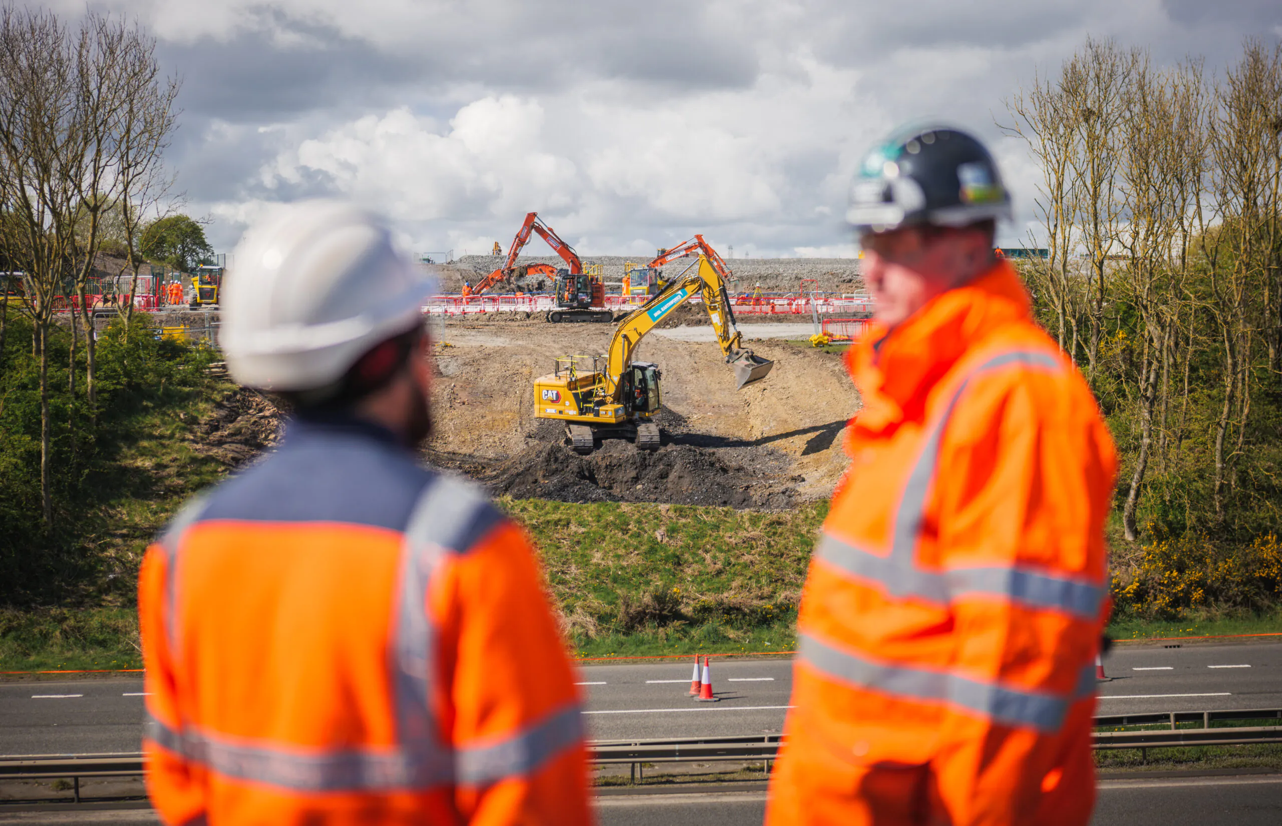 Two workers in orange high-visibility clothing and helmets observe excavation work by heavy machinery on a roadside.