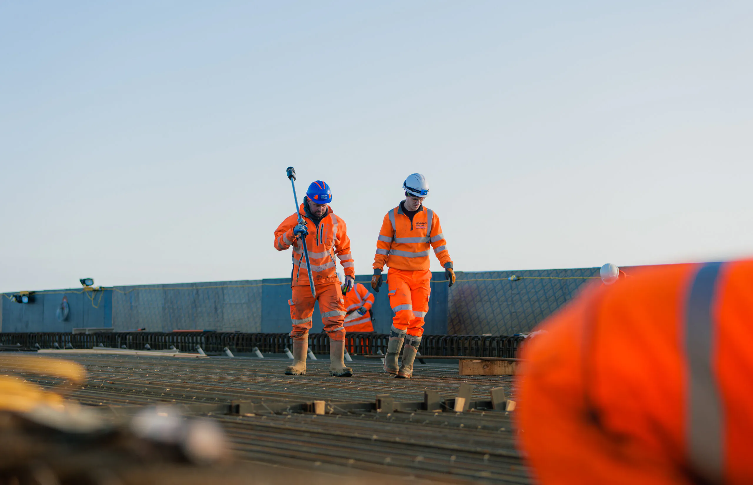 Two construction workers in orange safety gear walk across a site with metal structures and equipment; one carries a tool.