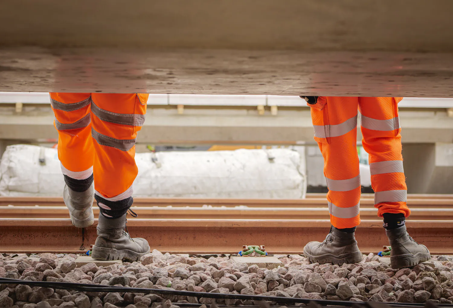 Lower legs of two workers in orange high-visibility trousers and work boots standing on railway tracks.