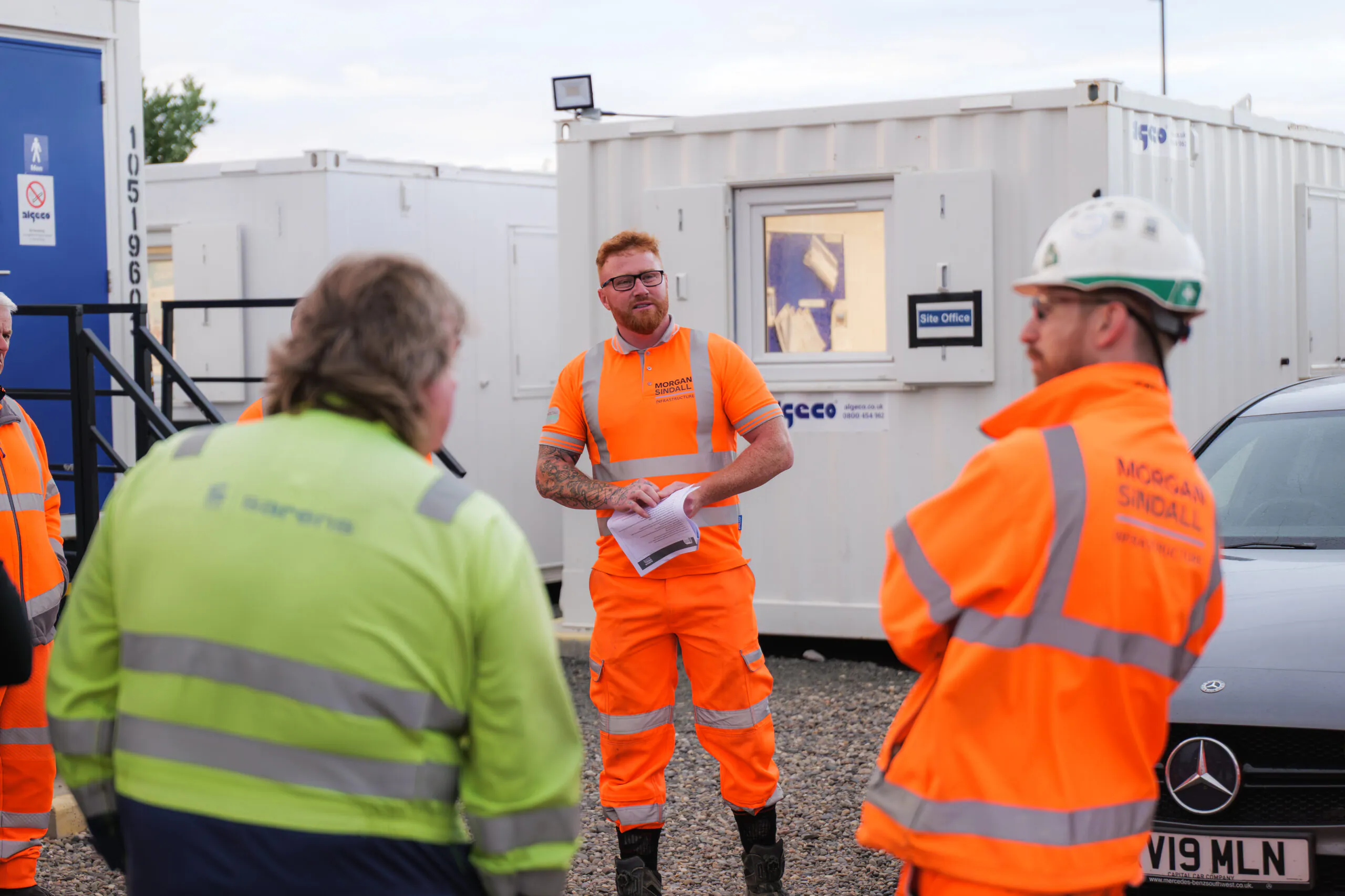 Worker in orange safety gear holds papers and addresses three others at a construction site with portable office units in the background.