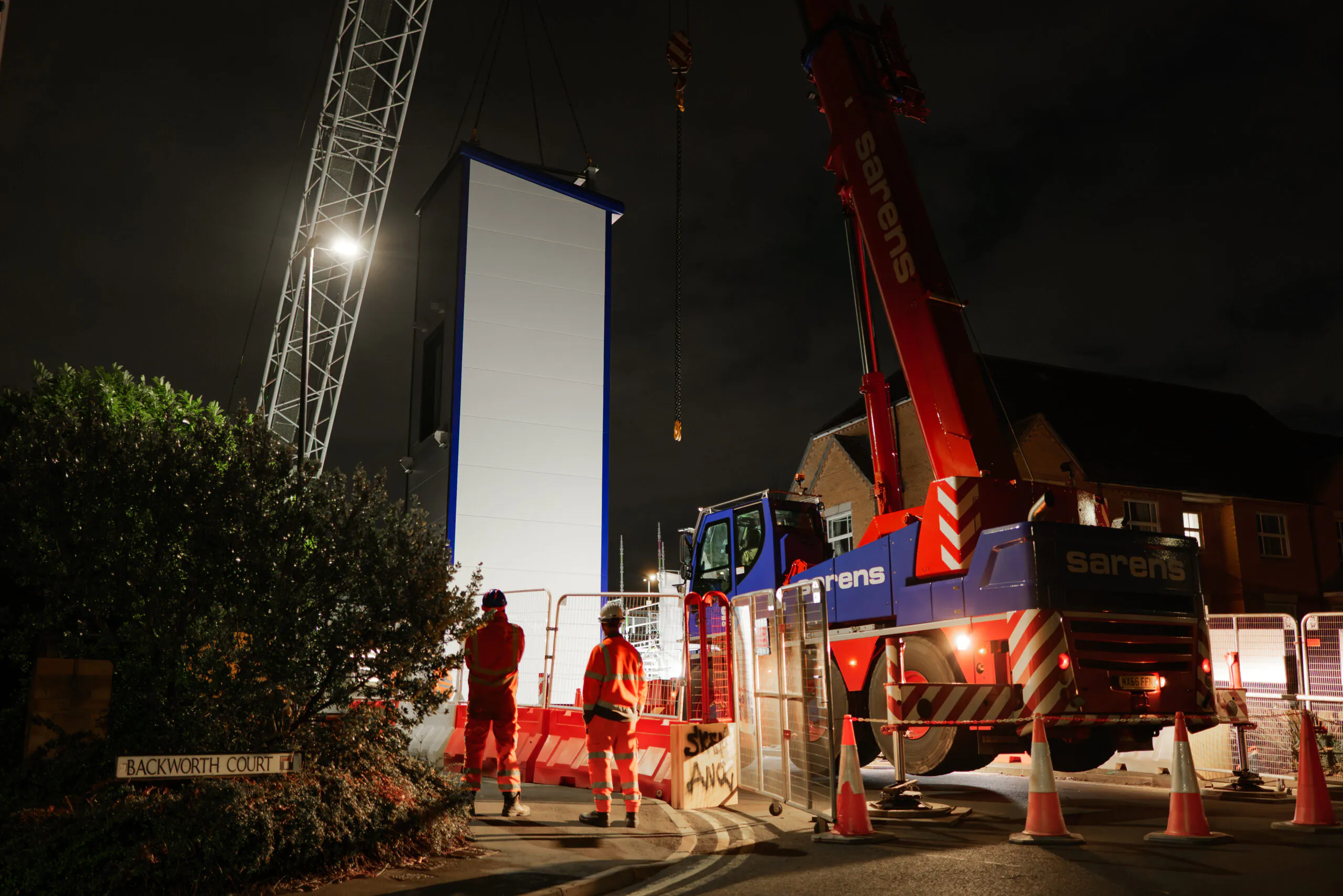 Nighttime construction scene with two workers in high-visibility gear near a crane lifting a tall structure, illuminated by floodlights.