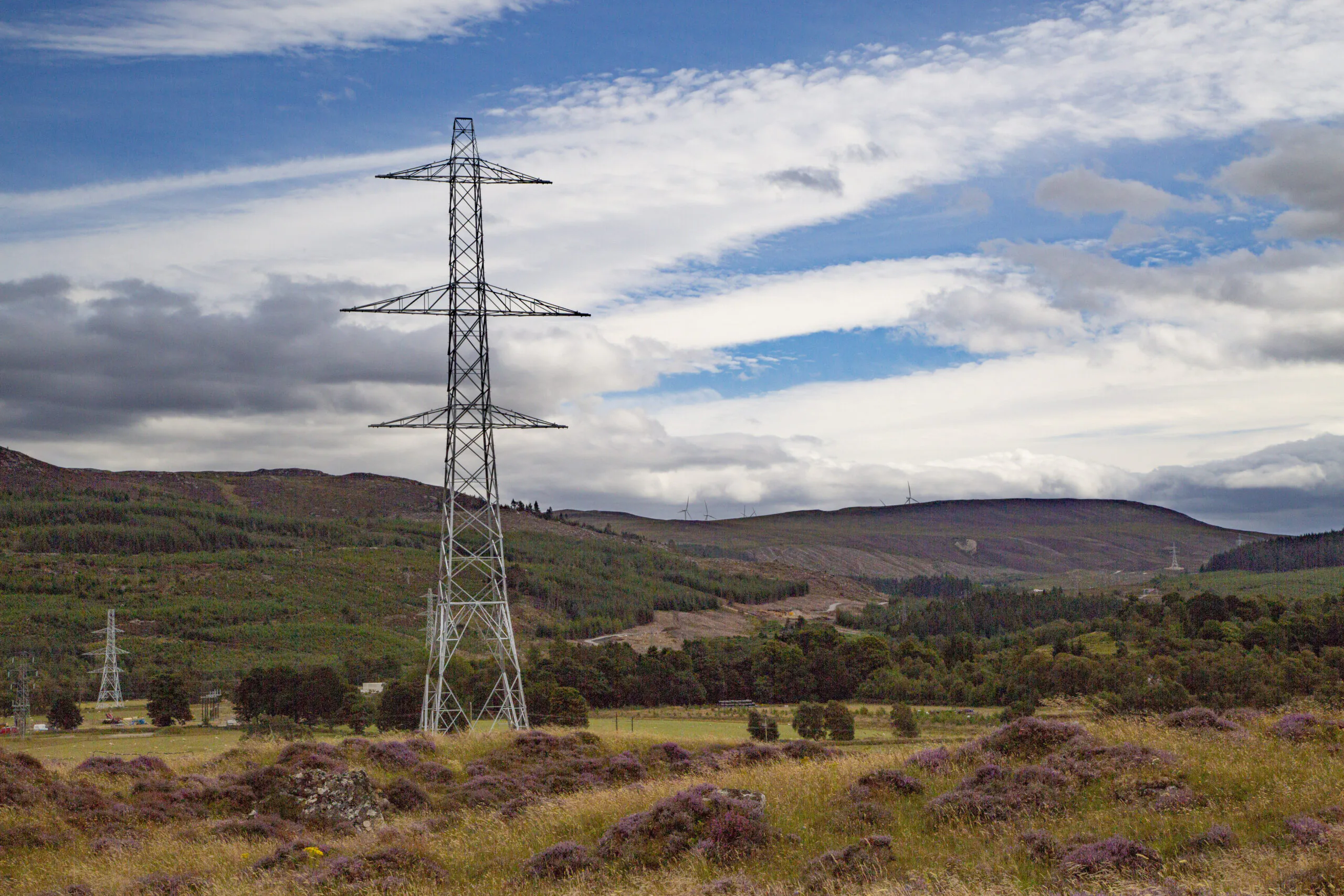 A tall electricity pylon stands in a scenic landscape of rolling green hills and purple heather, with another pylon visible in the distance under a partly cloudy sky.