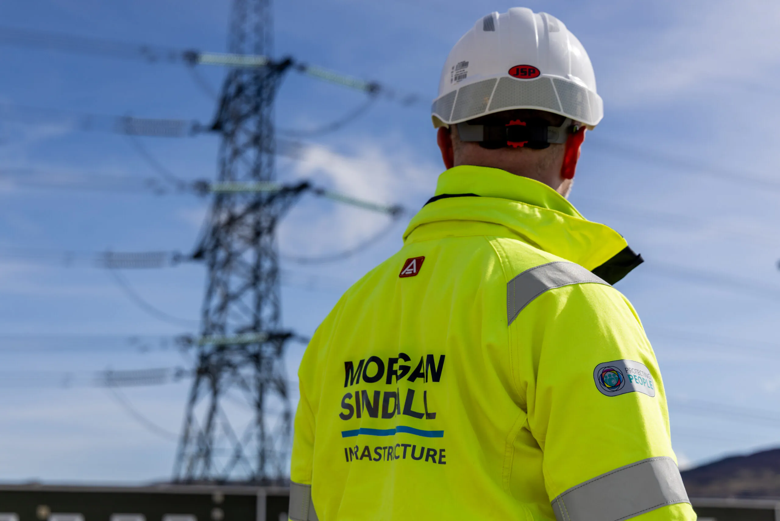 Person in a high-visibility jacket and hard hat stands facing large electrical pylons; the jacket reads “Morgan Sindall Infrastructure.”