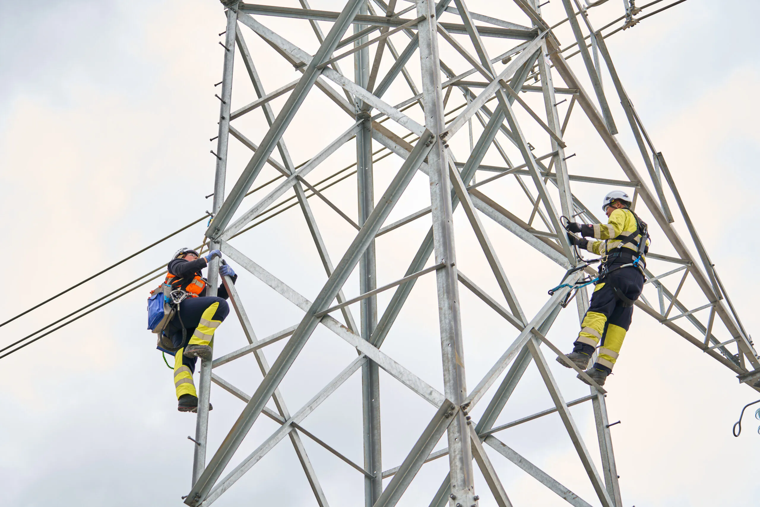 Two workers in safety gear climbing a metal lattice tower structure.