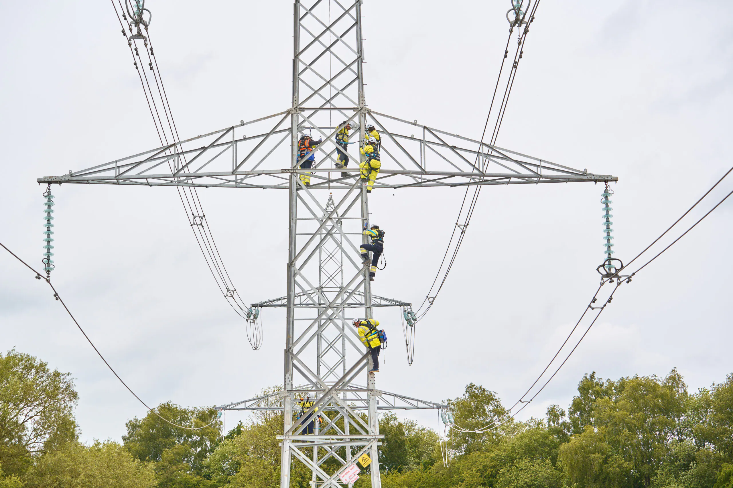 Workers in safety gear climbing and working on a tall metal lattice tower with power lines.