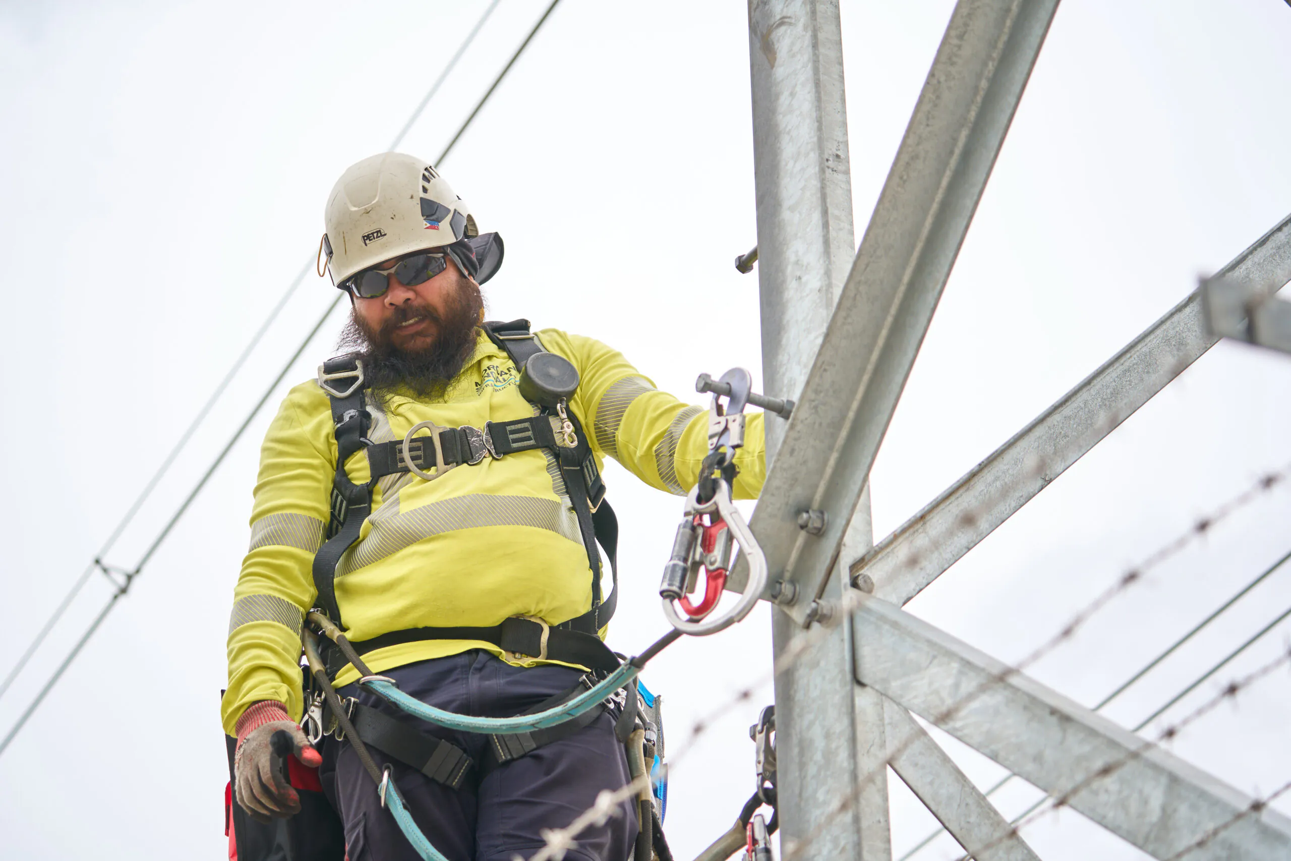 Worker at height wearing safety gear secures cables to metal structures against an overcast sky.