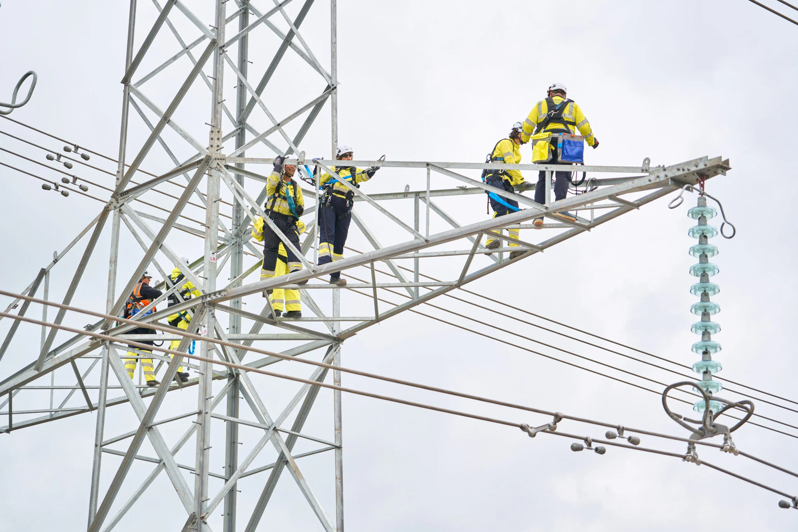 Multiple workers in high-visibility clothing working atop horizontal beams of a tall metal lattice tower with power lines.