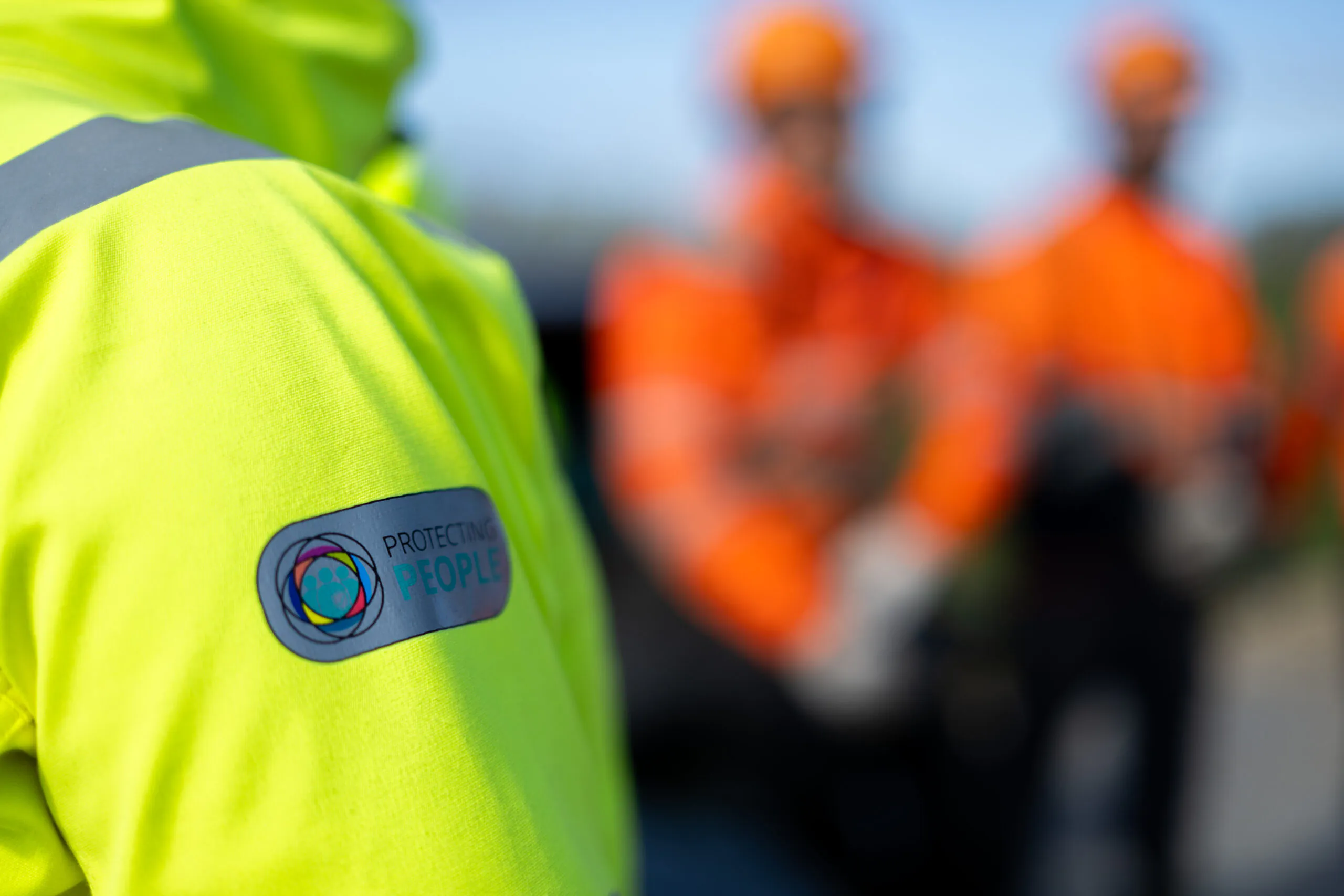 A close-up of a sleeve with the “Protecting People” logo patch, with blurred background.