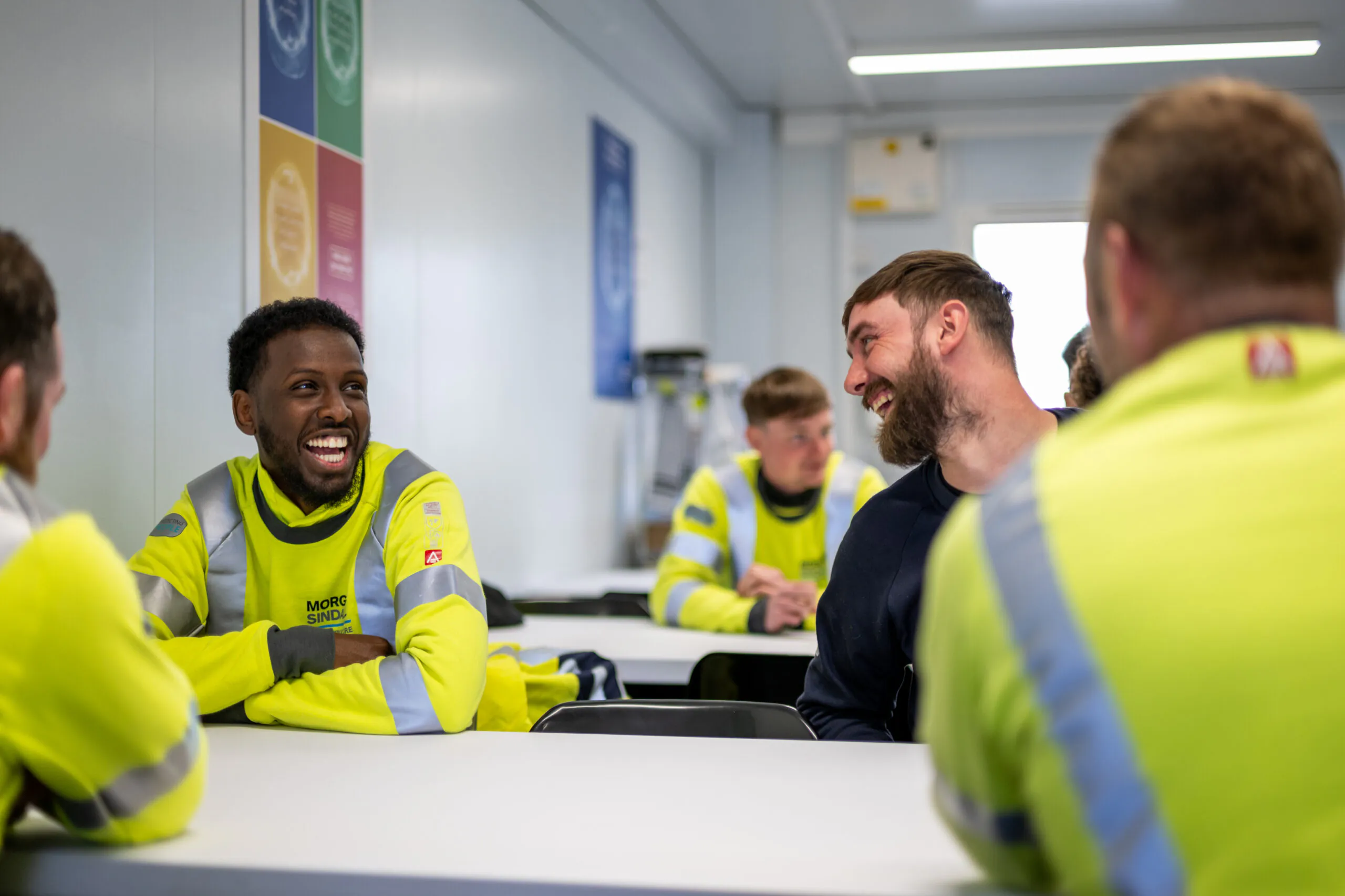 Group of workers in high-visibility clothing sit around tables in a break room, talking and smiling.