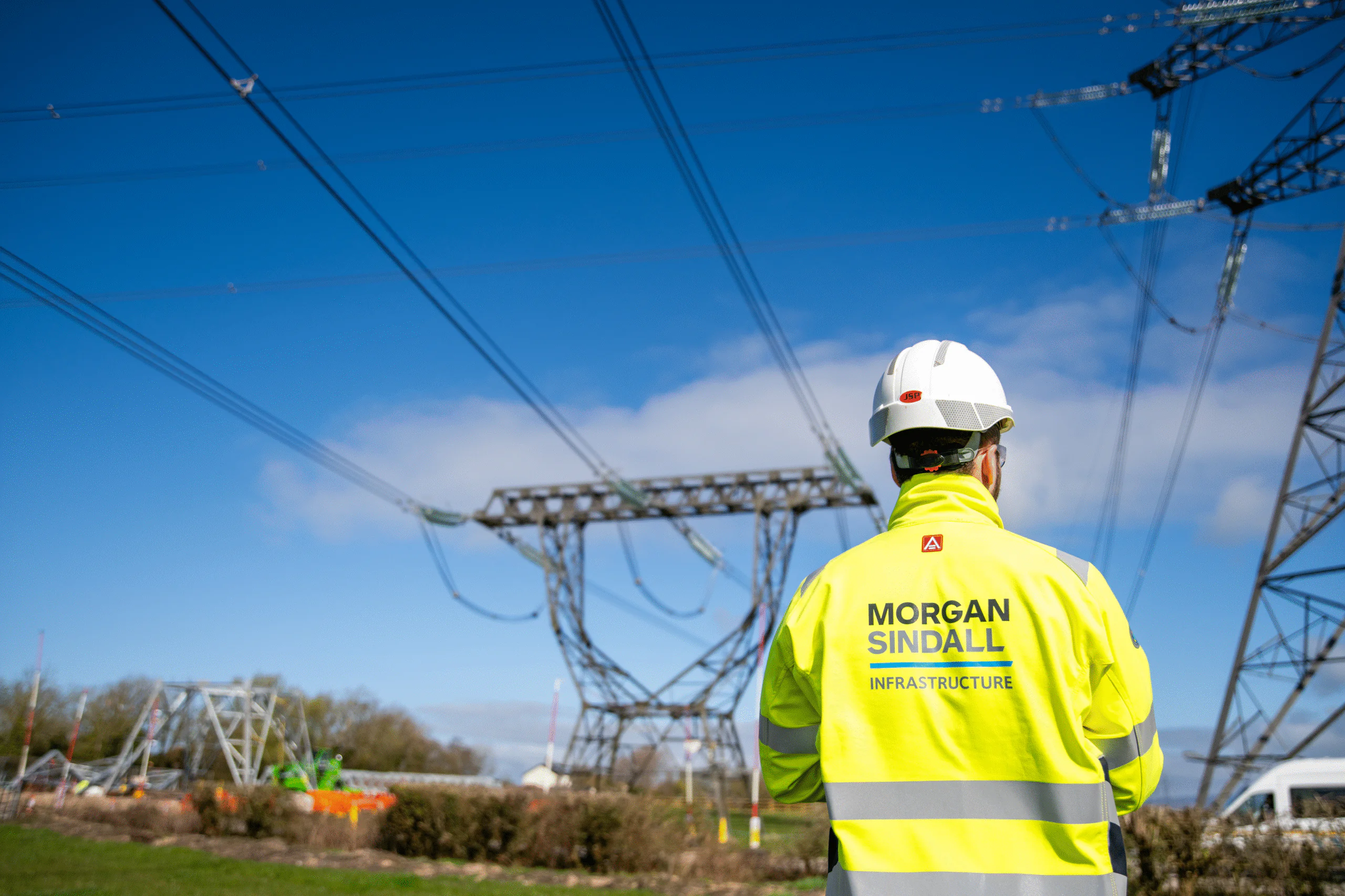 Person in a high-visibility jacket and hard hat stands facing large electrical pylons; the jacket reads “Morgan Sindall Infrastructure.”