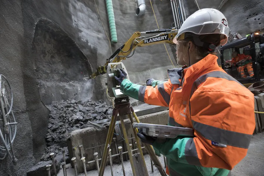 Worker operates surveying equipment inside an underground tunnel with rough rock walls; another worker operates machinery nearby.