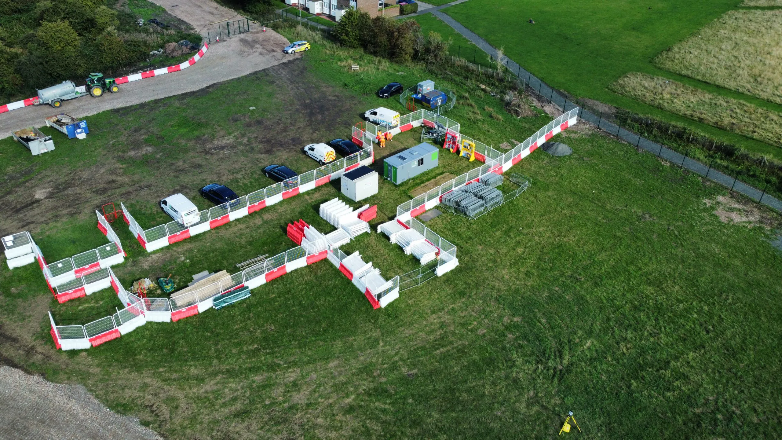 Aerial view of a fenced construction area with materials, equipment, and vehicles surrounded by grass fields and buildings.