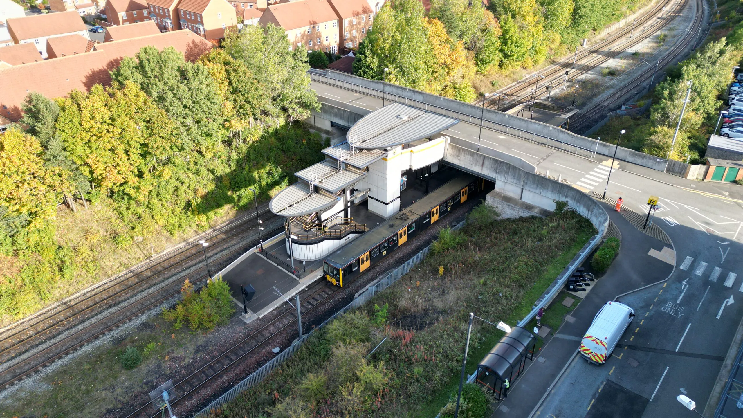Aerial view of a train station beneath a road bridge, with a train at the platform and surrounding trees and residential buildings.