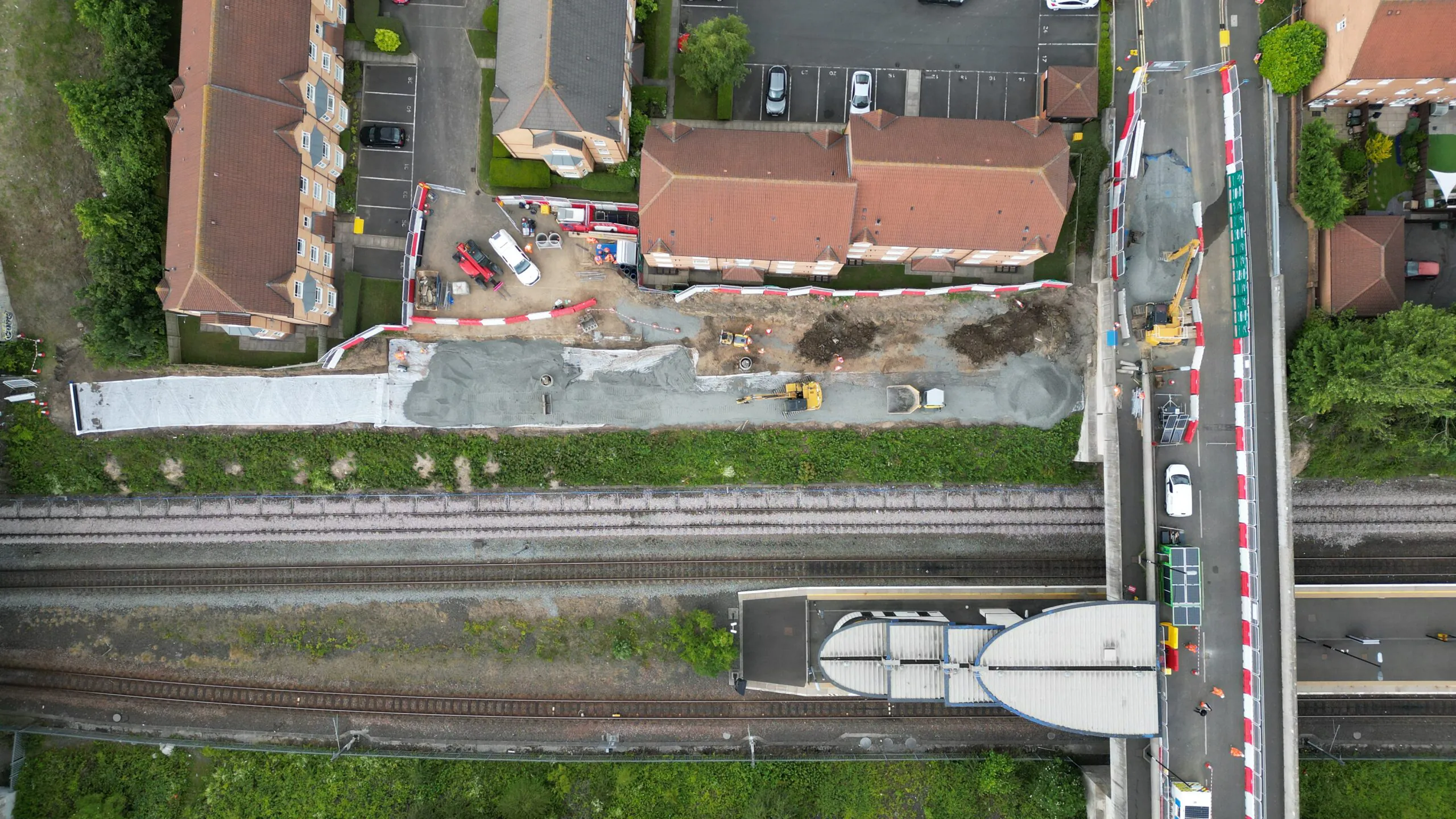 Overhead view of a construction site next to railway tracks and residential buildings, featuring construction vehicles and active workers.