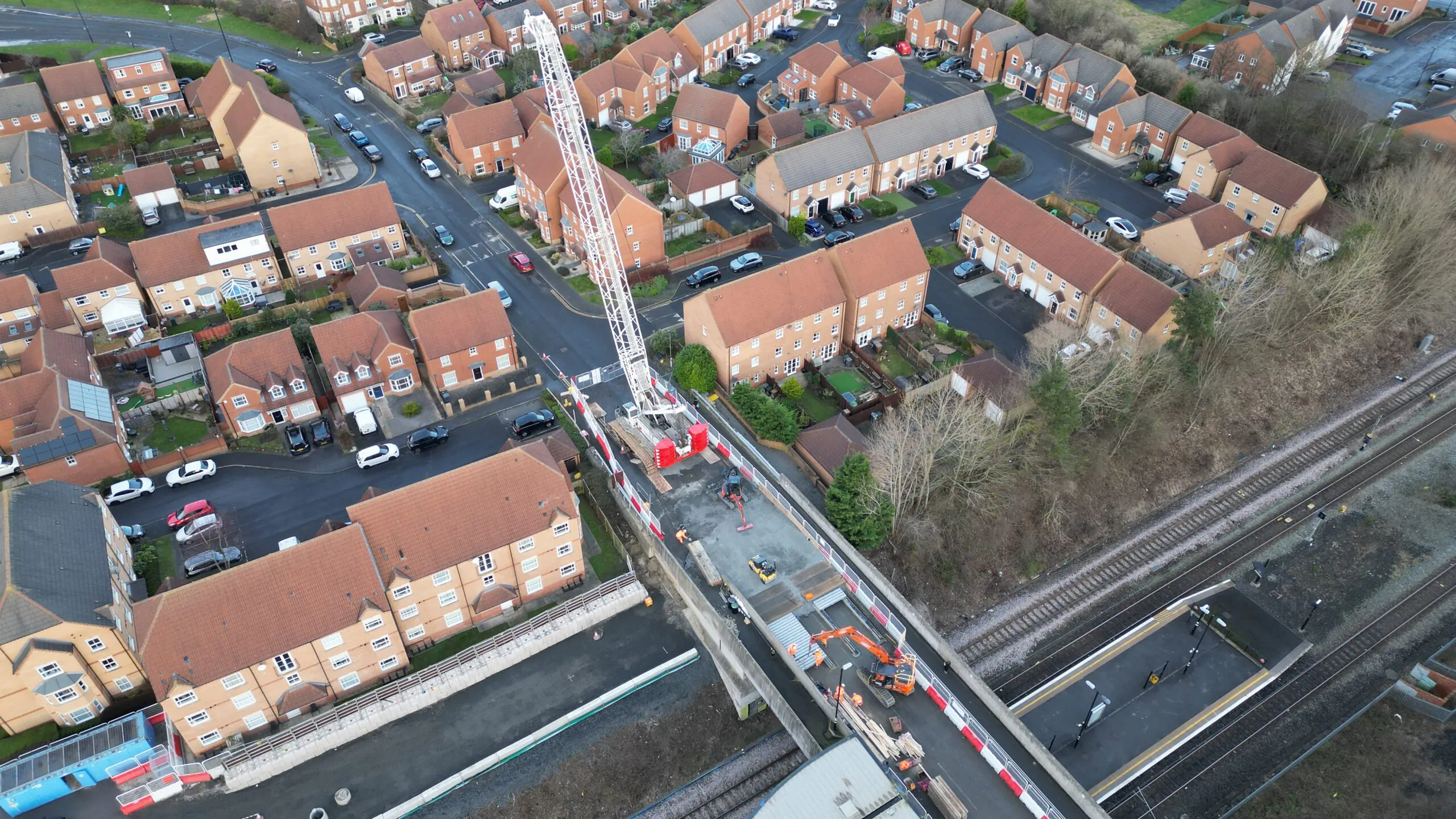 Wide aerial shot of a neighborhood with houses surrounding a railway-side construction site, where a large crane is positioned over a bridge.