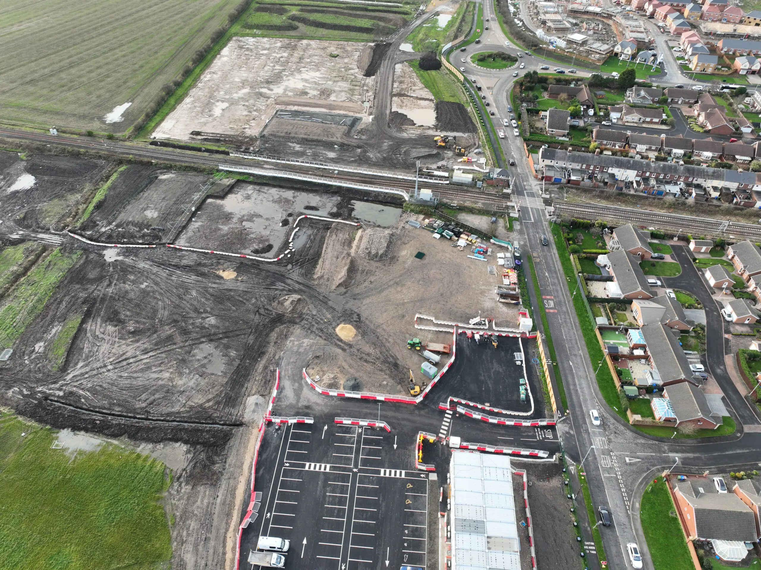 Aerial view of a large construction site near residential areas and fields, with excavated zones and temporary structures.