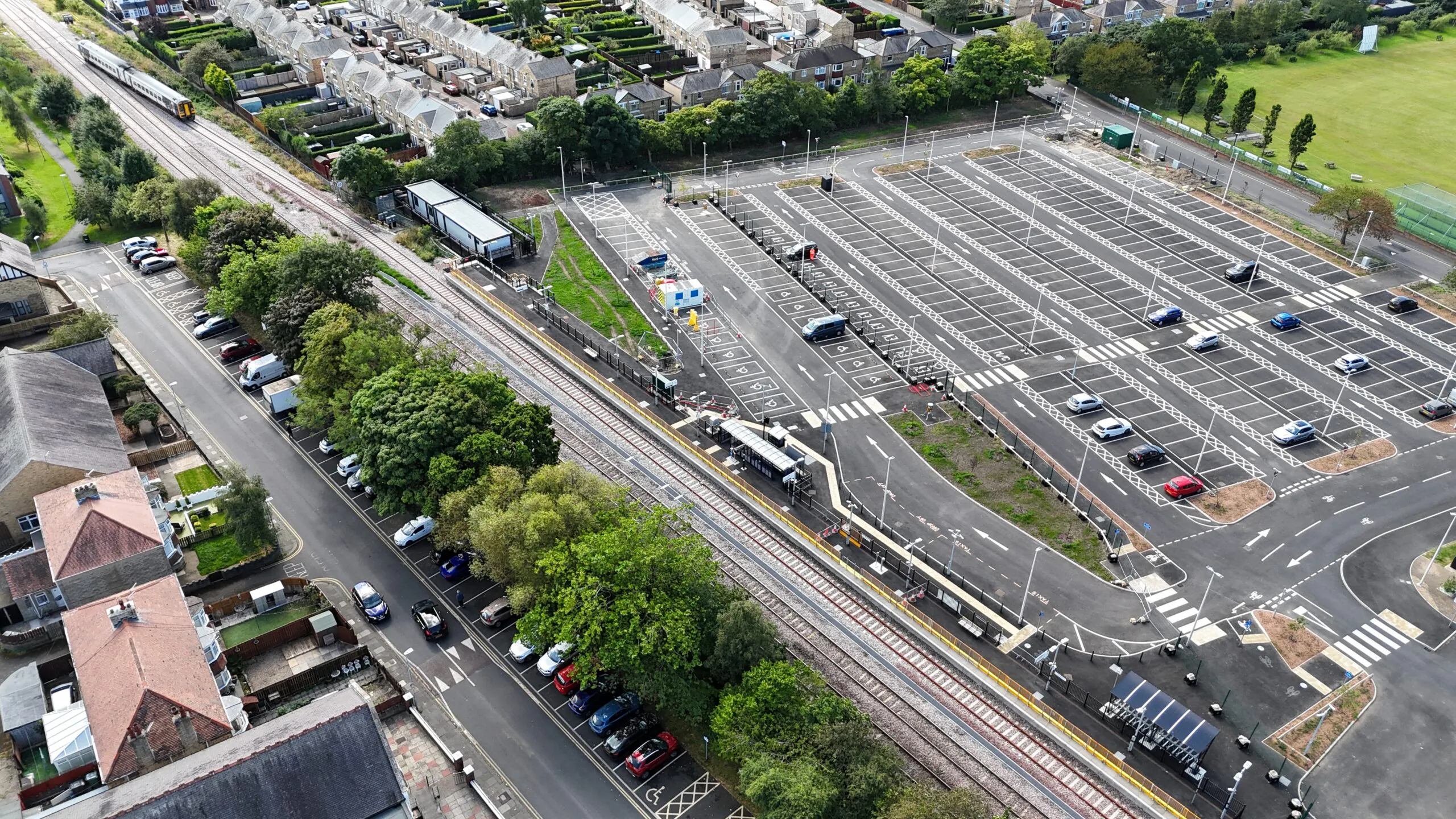 Aerial view of an empty parking lot adjacent to railway tracks, surrounded by greenery and buildings.