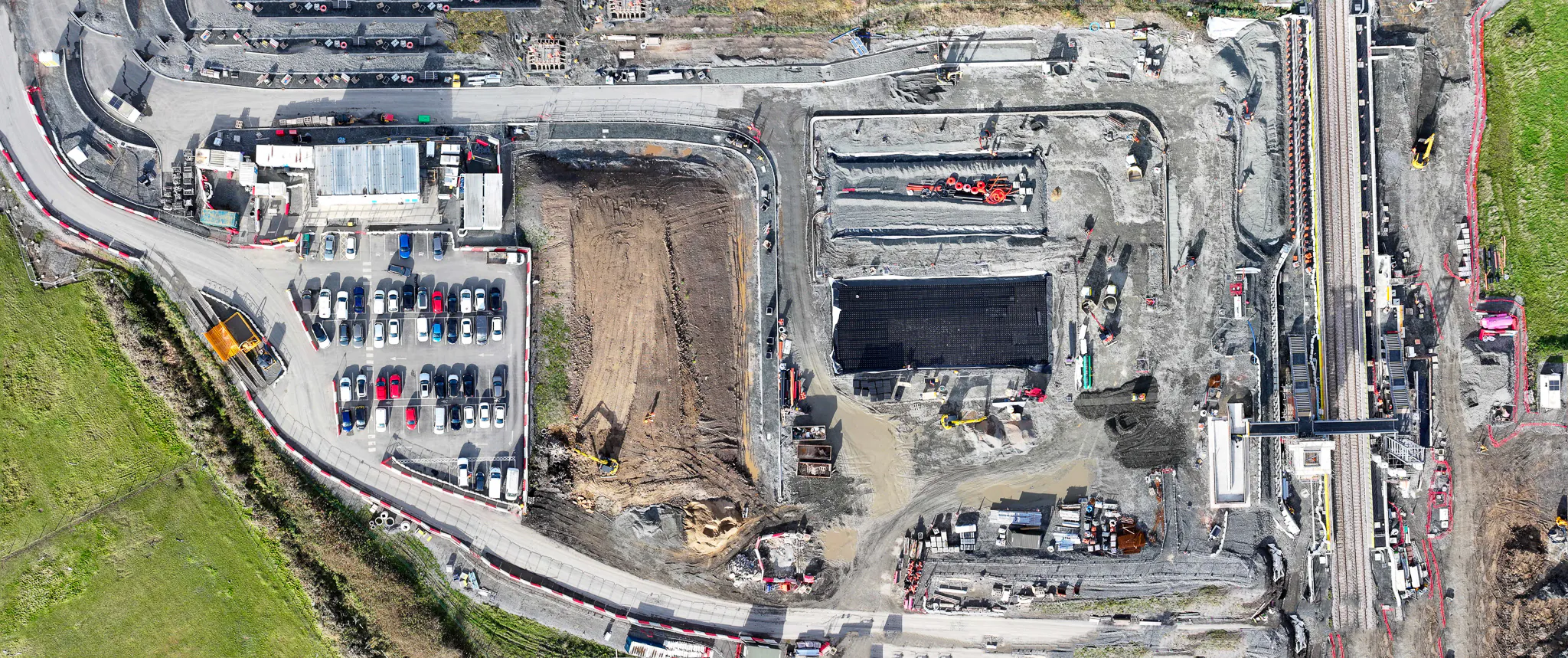 Overhead shot of excavation work within fenced boundaries, surrounded by green fields and parked vehicles.