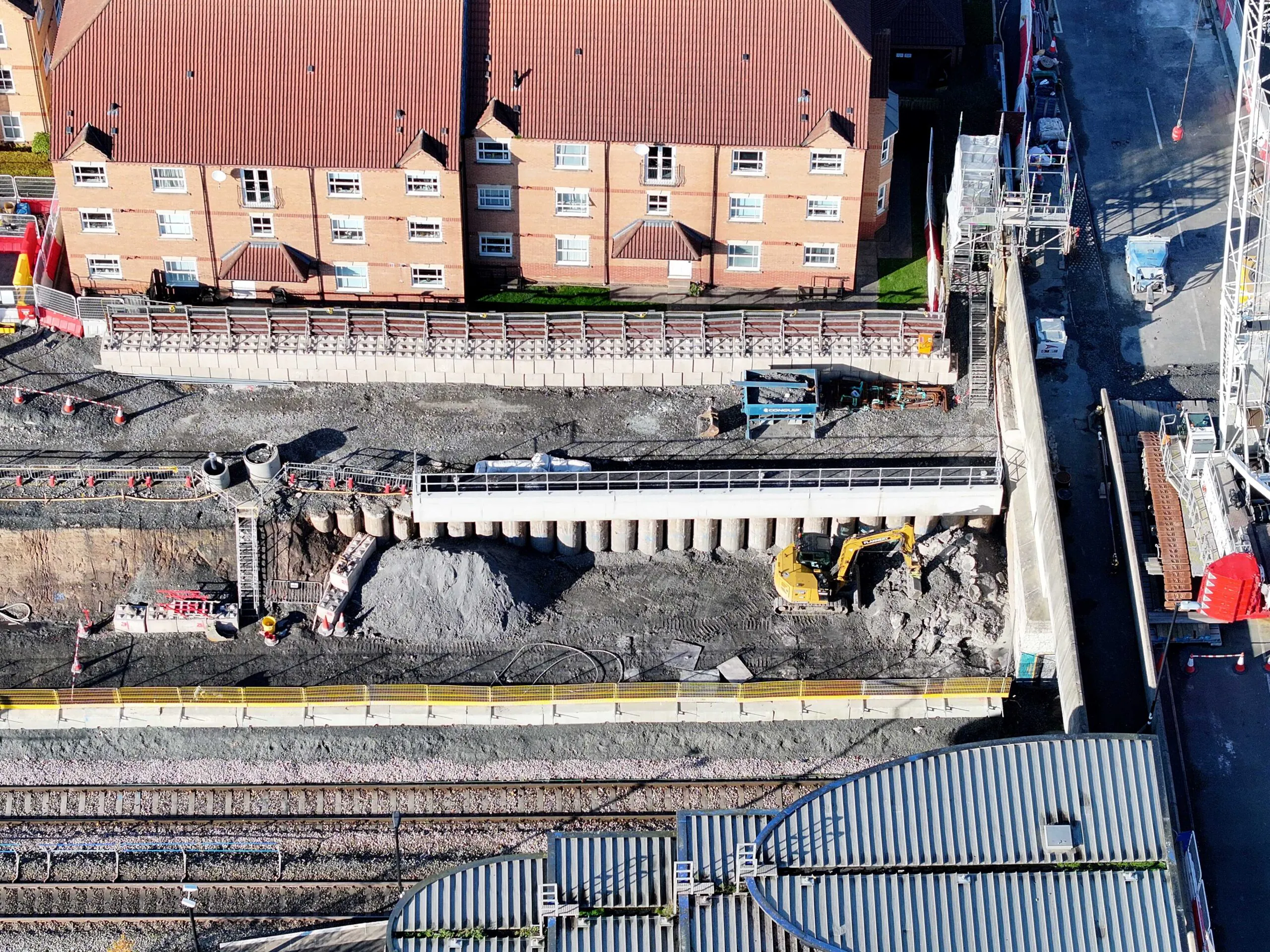 Aerial view of a construction site near railway tracks with residential buildings in the background and visible construction materials.