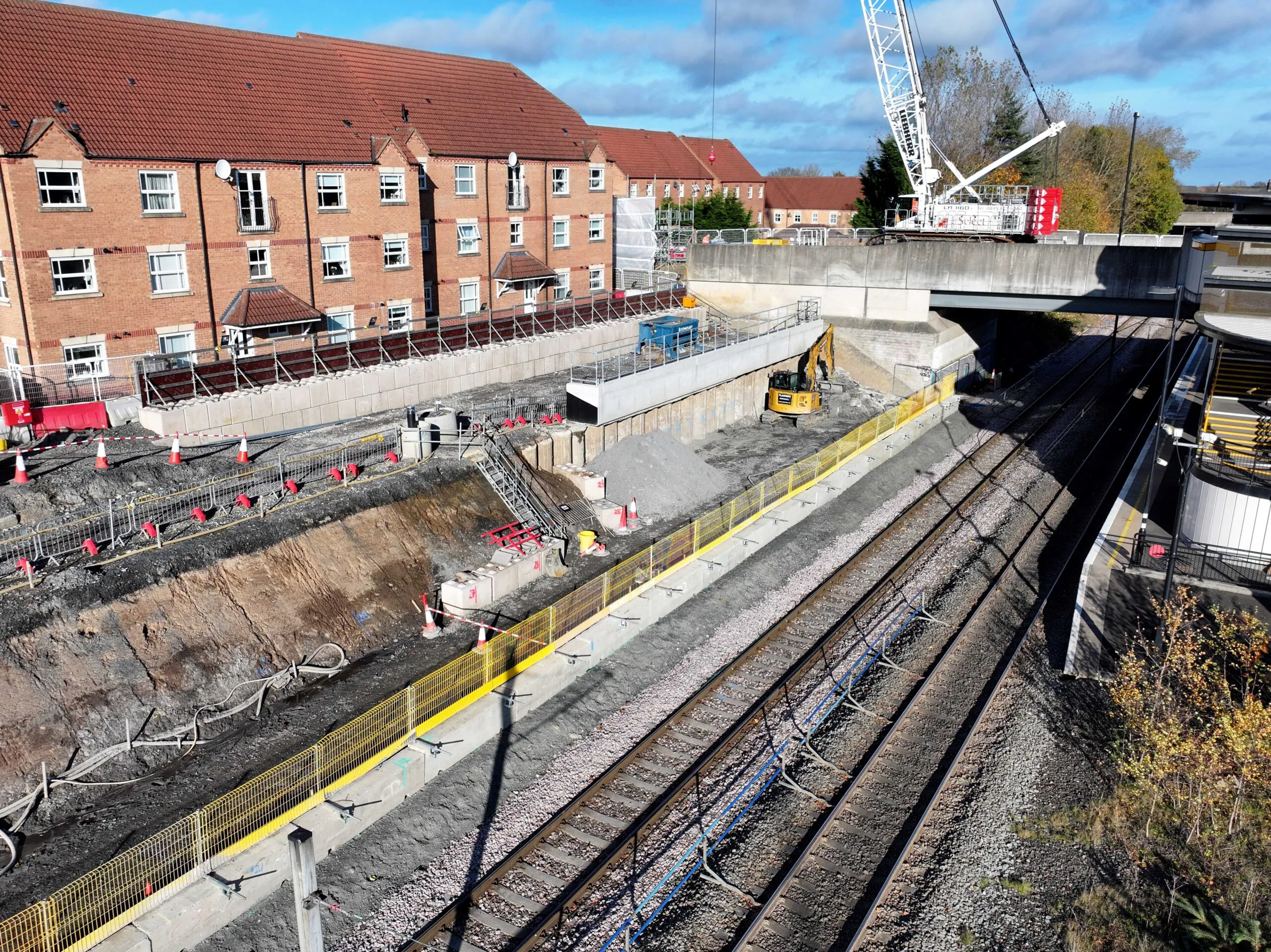 Aerial view of a railway-side construction site with heavy machinery including a crane and excavator, adjacent to residential buildings with red roofs.