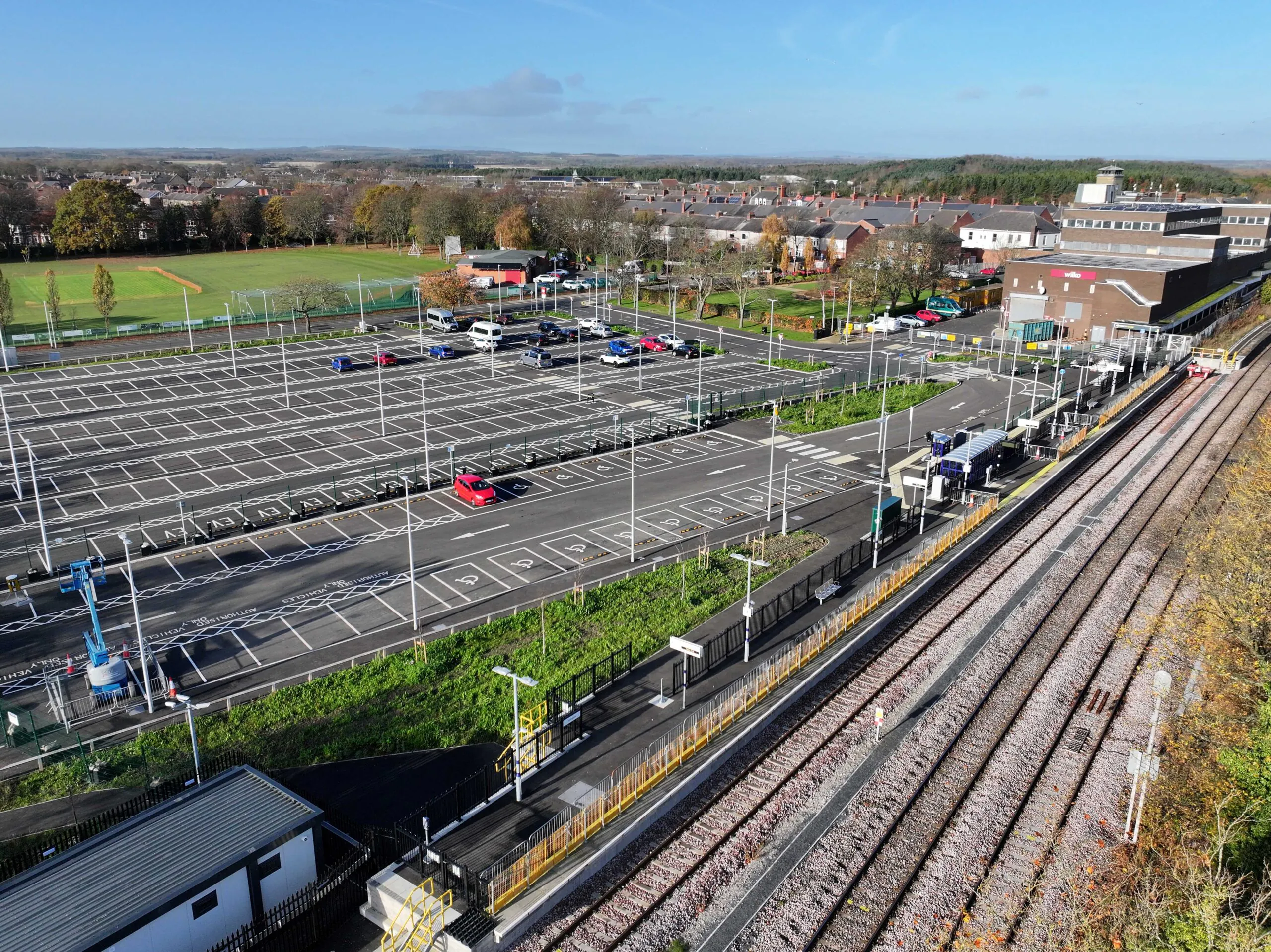 Aerial view of an empty parking lot adjacent to railway tracks, surrounded by greenery and buildings.