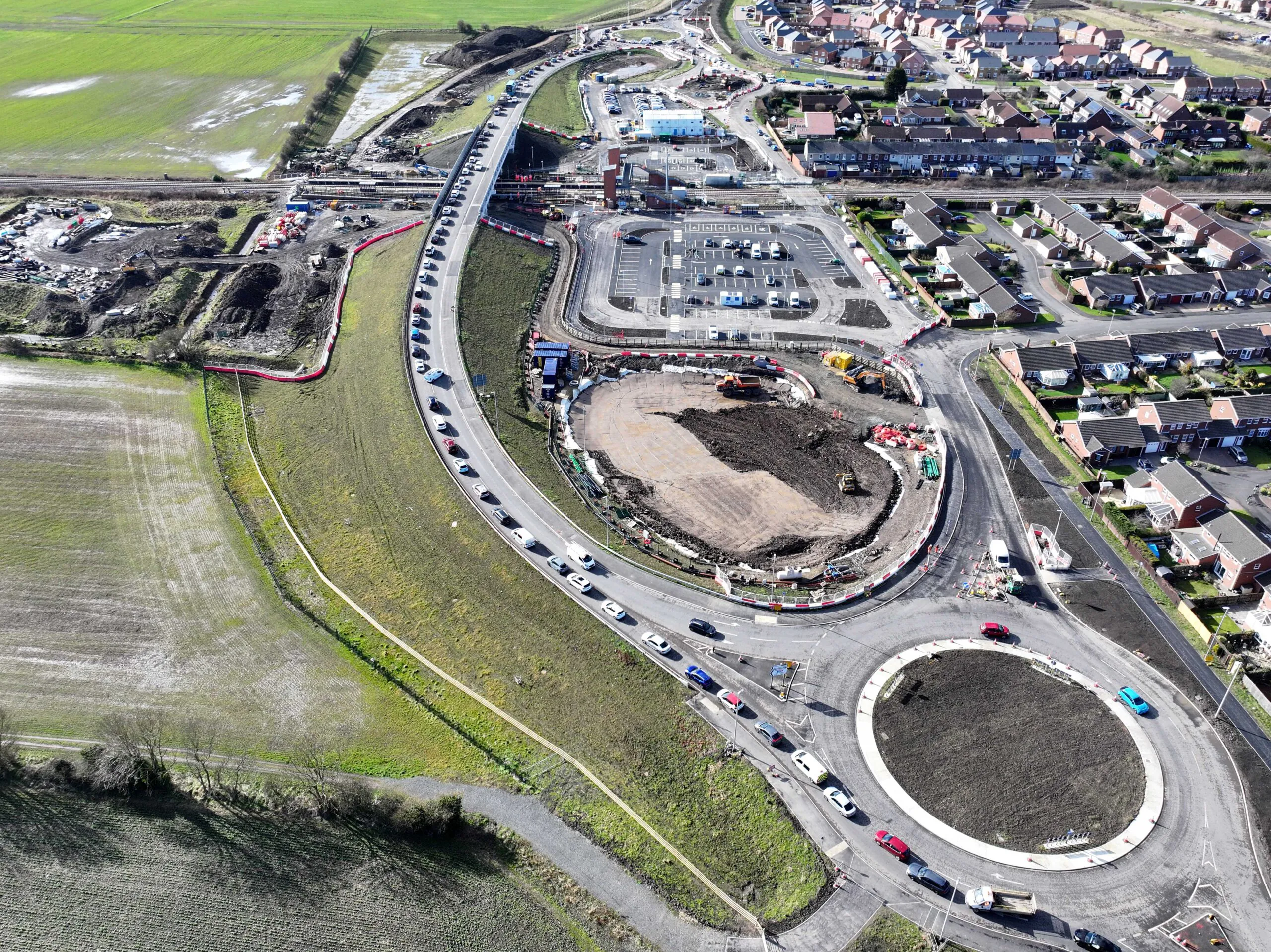 Aerial view of extensive roadwork with roundabouts, parking lots, and temporary structures near residential areas.