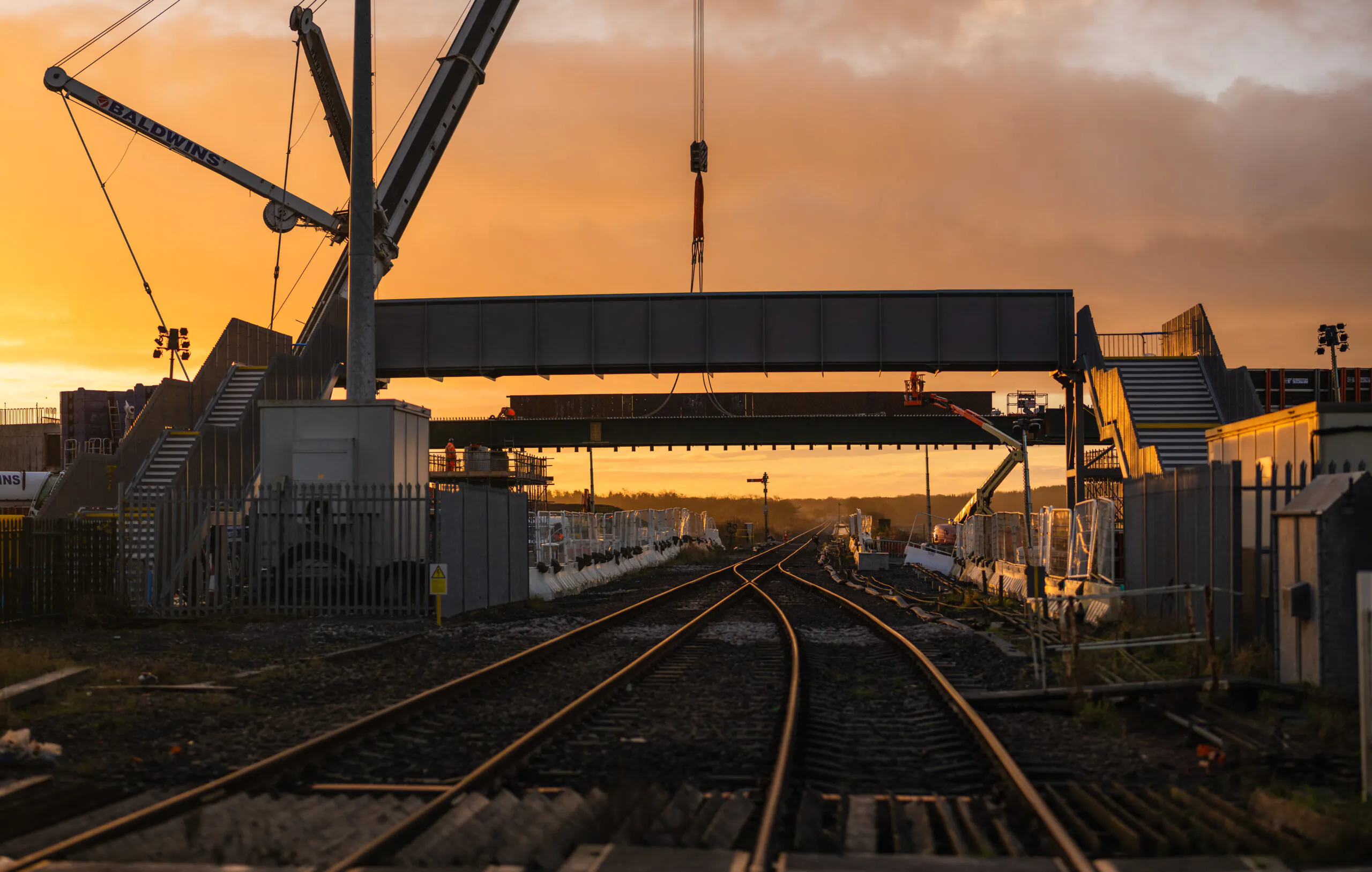 Sunrise view of railway tracks leading to a pedestrian bridge being lifted into place by cranes.