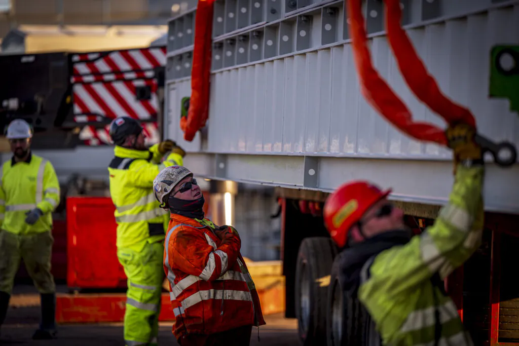 Red crane lifts structural components at a construction site while workers observe from below.