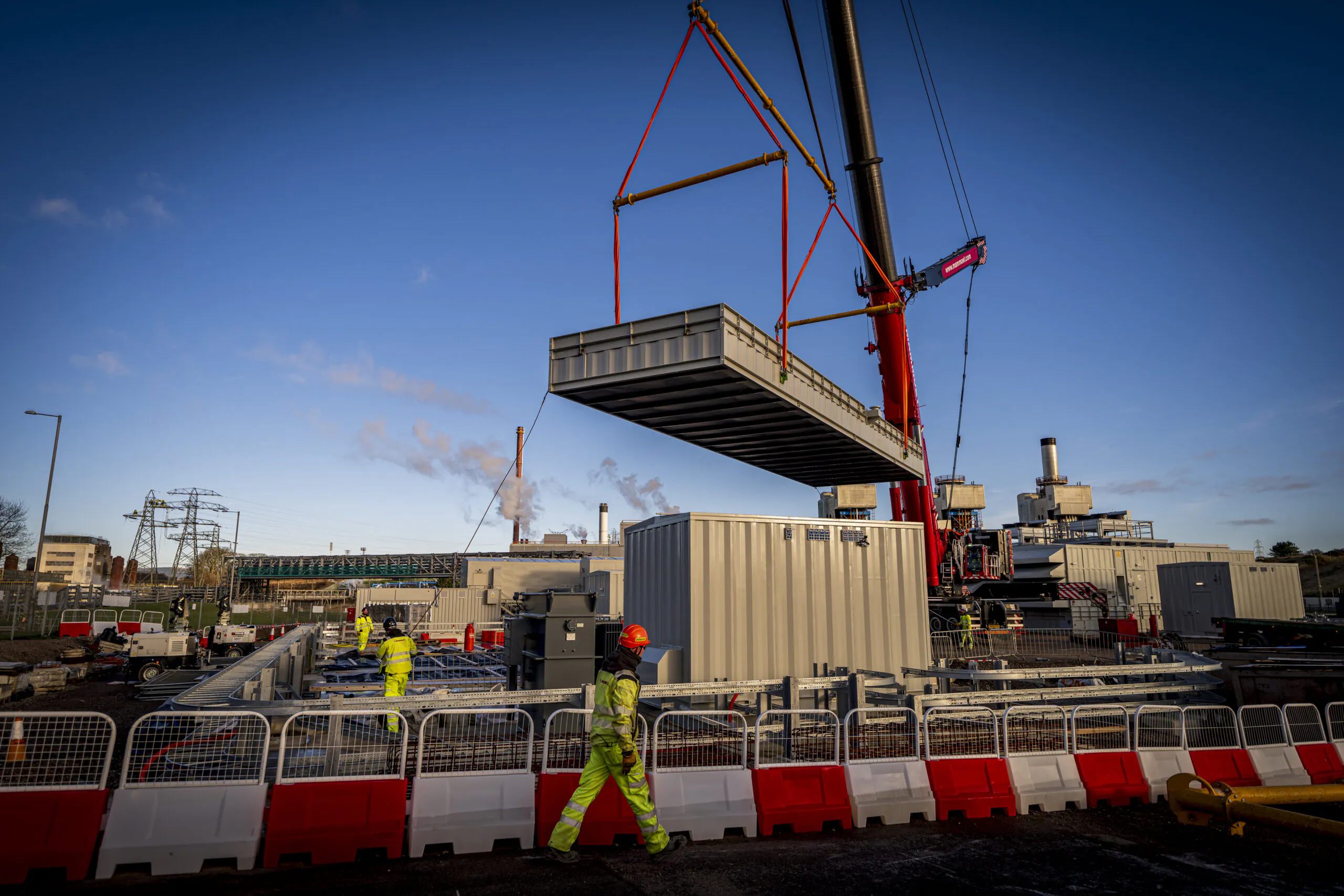 Large red crane lifts part of a building structure while workers observe below; safety barriers surround the area.