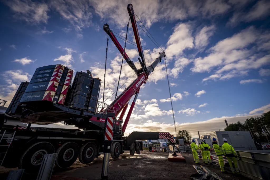 Large red crane lifts heavy materials at a construction site with workers observing below under a partly cloudy sky.
