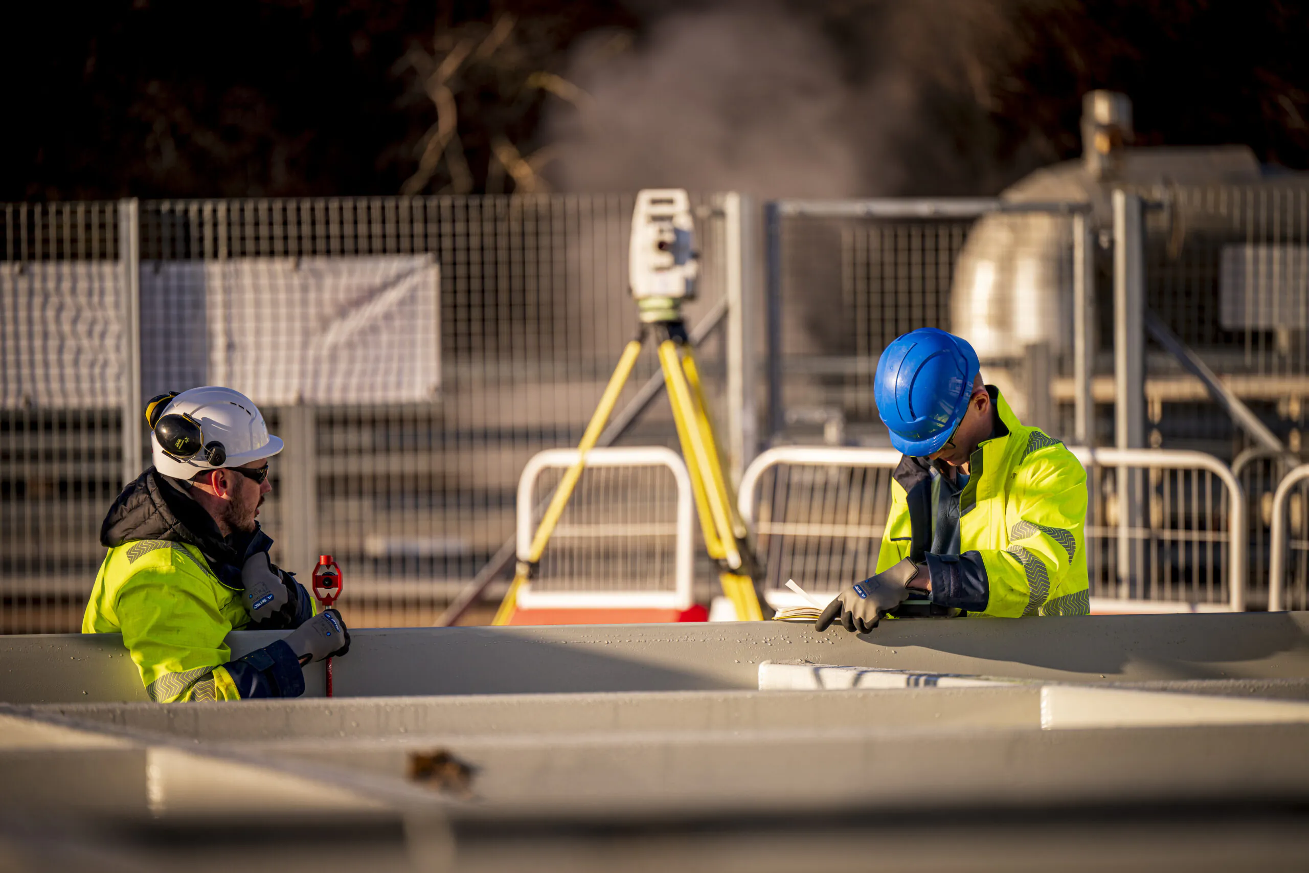 Two construction workers in high-visibility clothing work on-site, one holding a tool while the other inspects a structure.